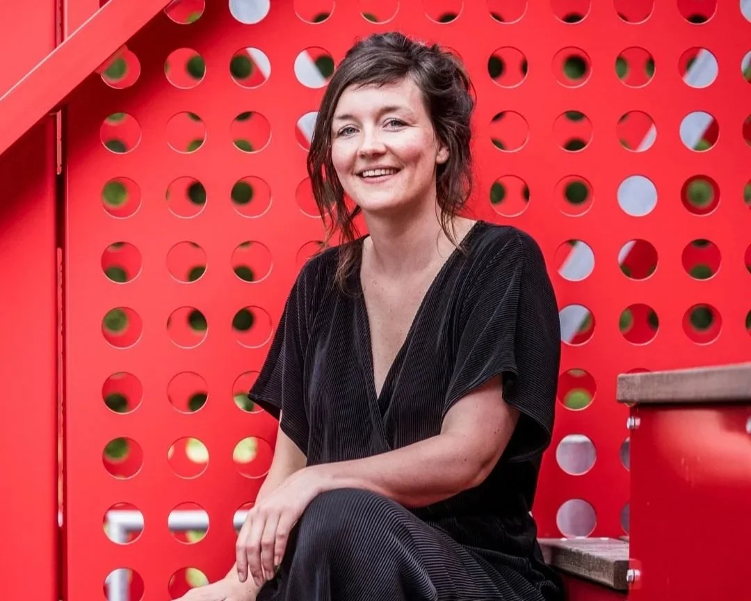 Een lachende vrouw met donkerbruin haar, zittend op een houten bankje voor een rood metalen hek met ronde gaten.