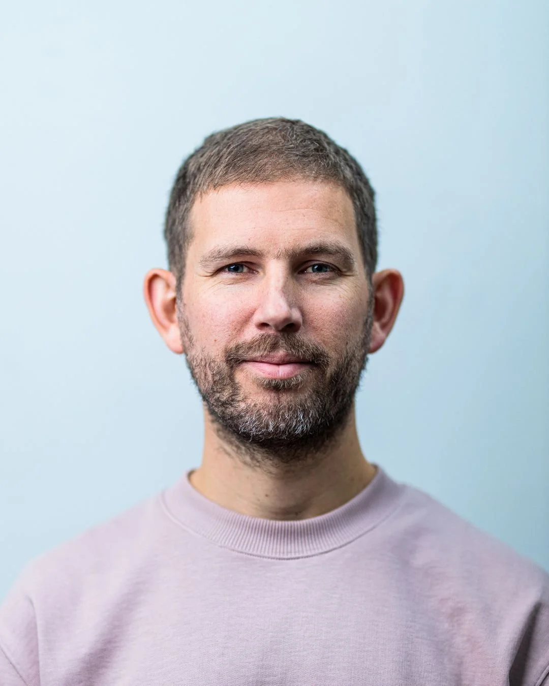 Portret van een man met kort bruin haar en een baard, gekleed in een lichte roze trui, staande voor een lichtblauwe achtergrond.