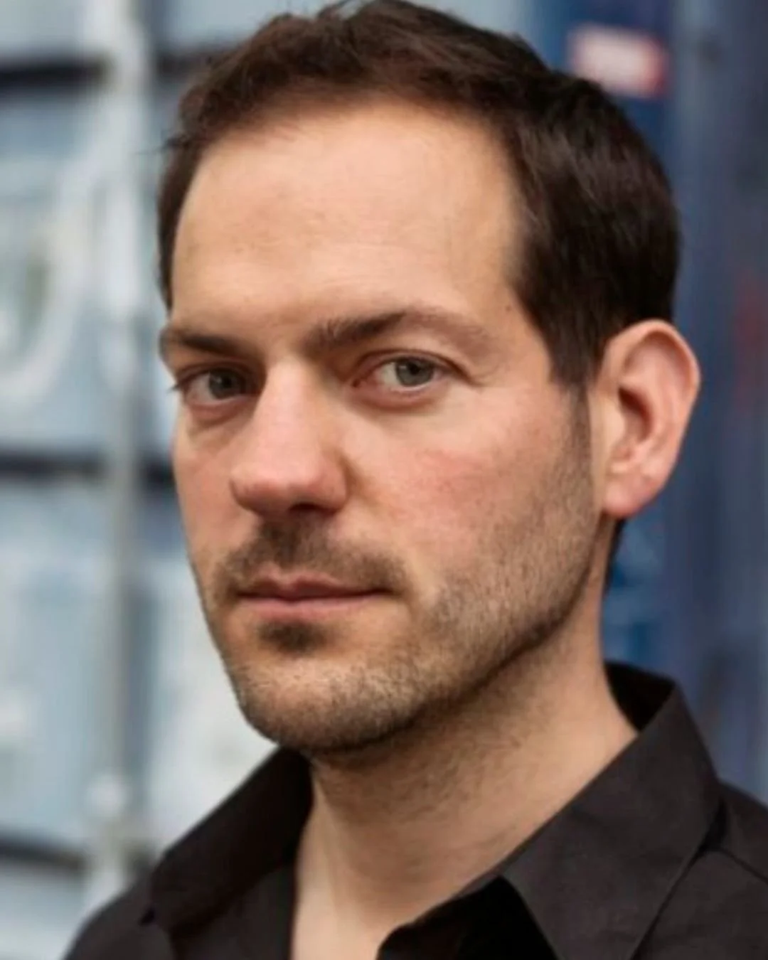 Close-up van een man met kort bruin haar en een zwart shirt, met een neutrale uitdrukking, op de achtergrond een metalen structuur.