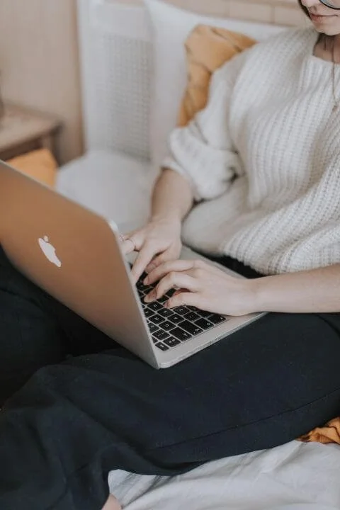 Person typing on a laptop at a white desk, wearing a cozy cream-colored sweater. There is a stack of books and a notebook in front of the person, with a large white vase filled with dried pampas grass and other dried flowers in the background.