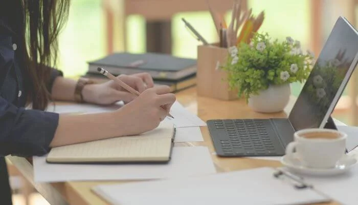 woman working outside on wooden desk with books, notebook, and laptop surrounding her.
