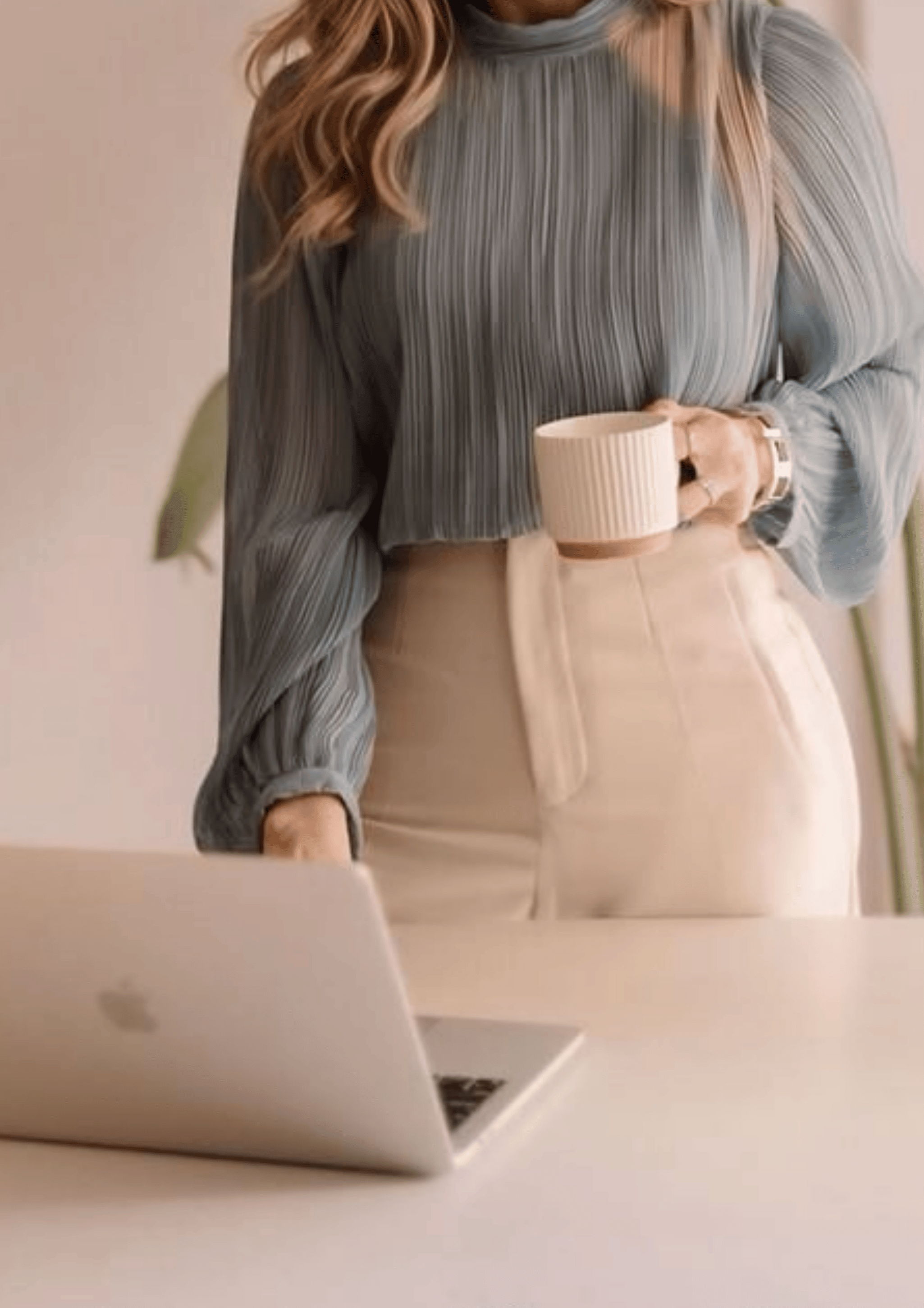 woman in white pants and blue blouse  standing at a desk with laptop open and coffee cup in hand.