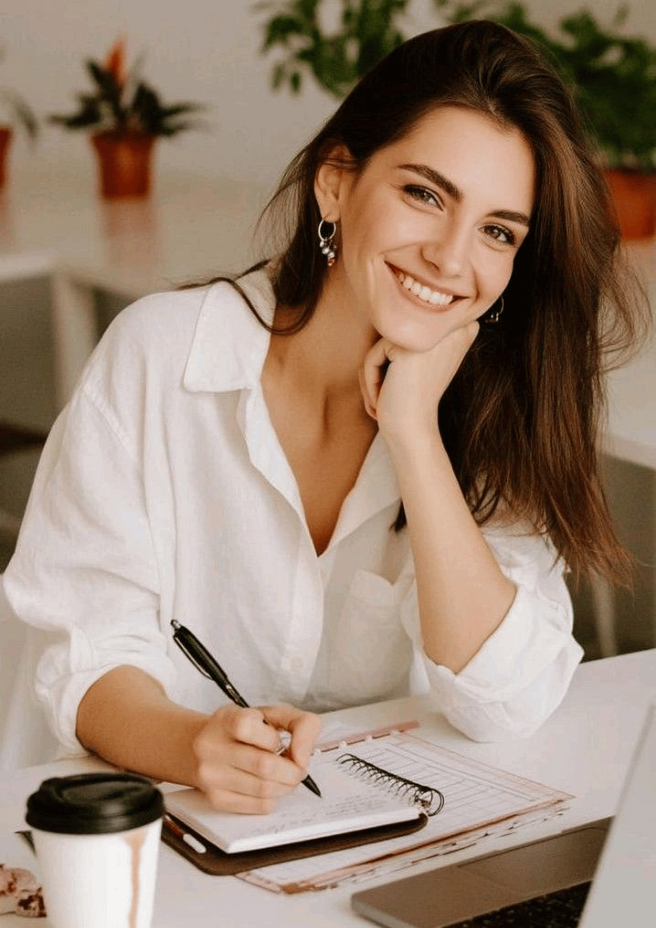 dark haired women in white blouse sitting at desk looking up smiling while pen in hand writing in day planner.