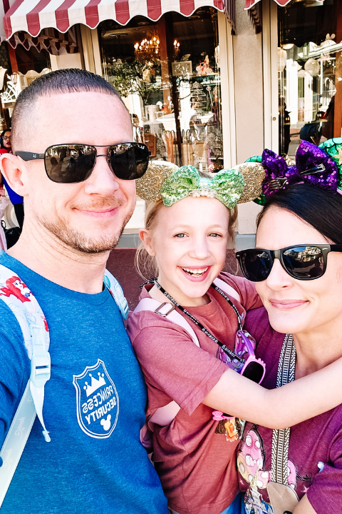 A smiling family of three posing for a photo outdoors, with a storefront window in the background. The man and woman are wearing sunglasses, and the girl has a colorful bow headband.