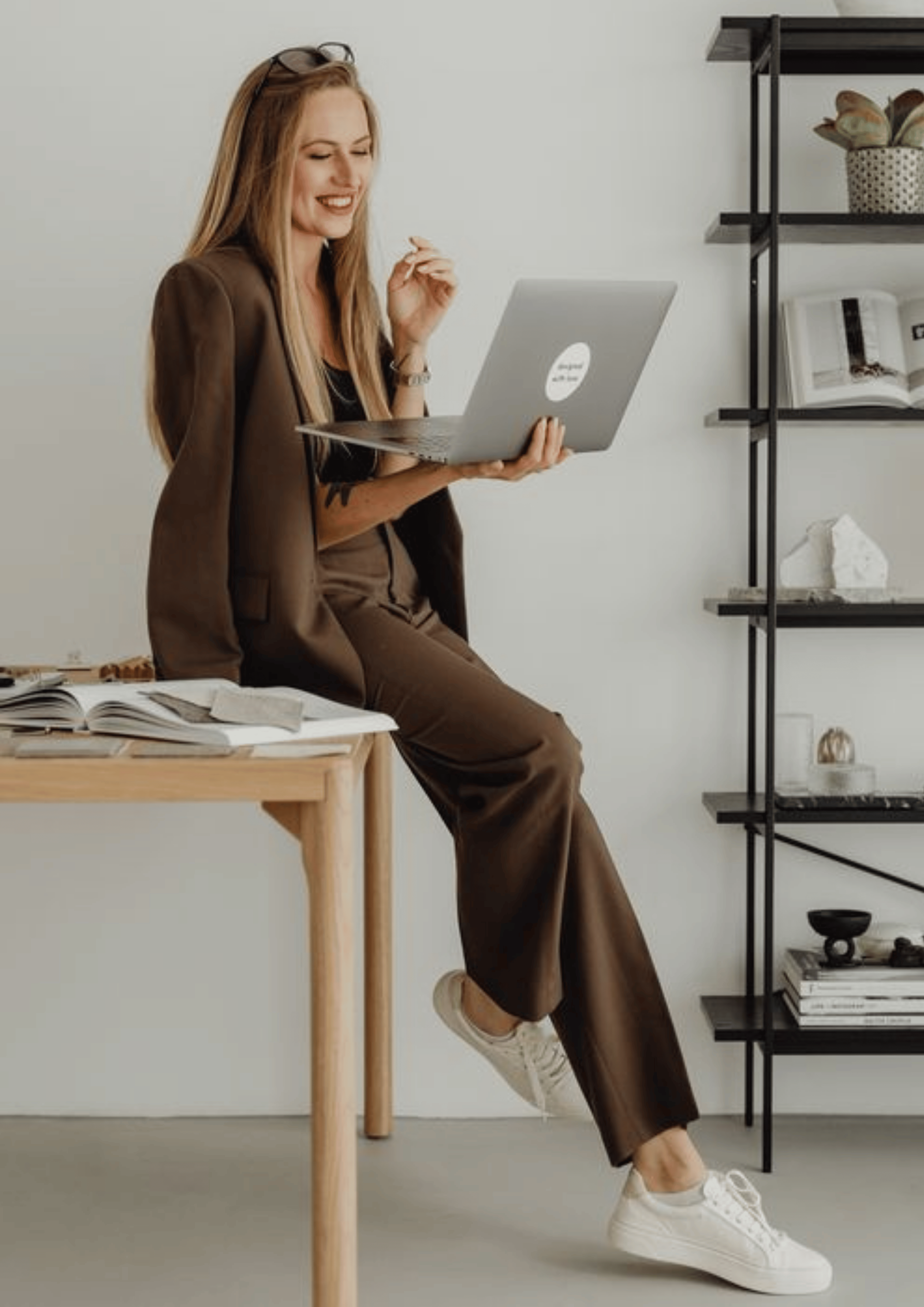 woman in brown pant suit and white sneakers sitting on edge of desk with her laptop in hand.