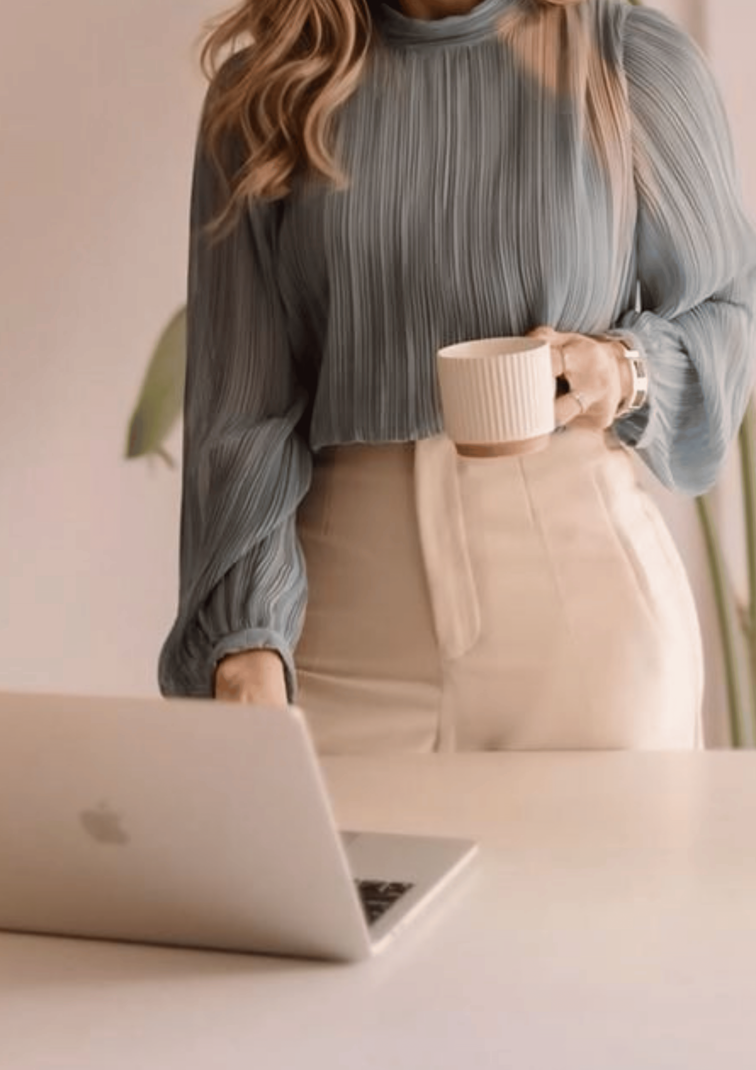 A woman is holding a beige mug, standing by a desk with a laptop, wearing a gray pleated blouse and beige pants.