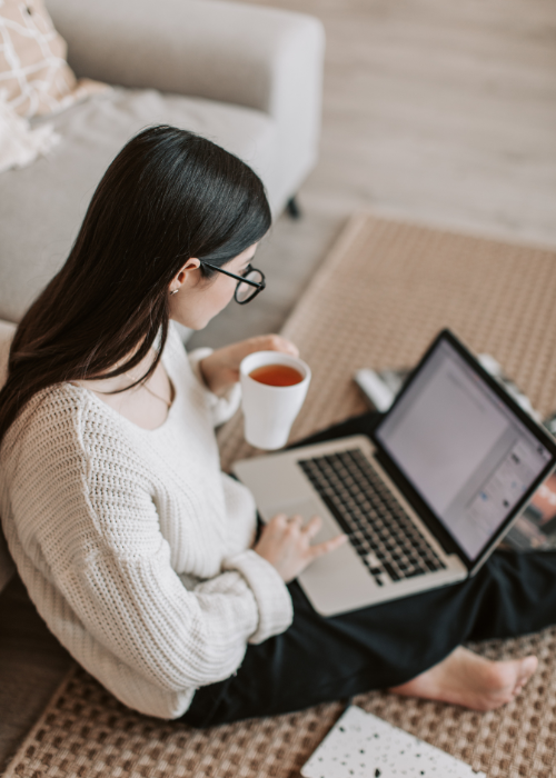 Woman sitting on carpeted floor with a laptop on her lap, holding a white mug, wearing glasses and a cream sweater.
