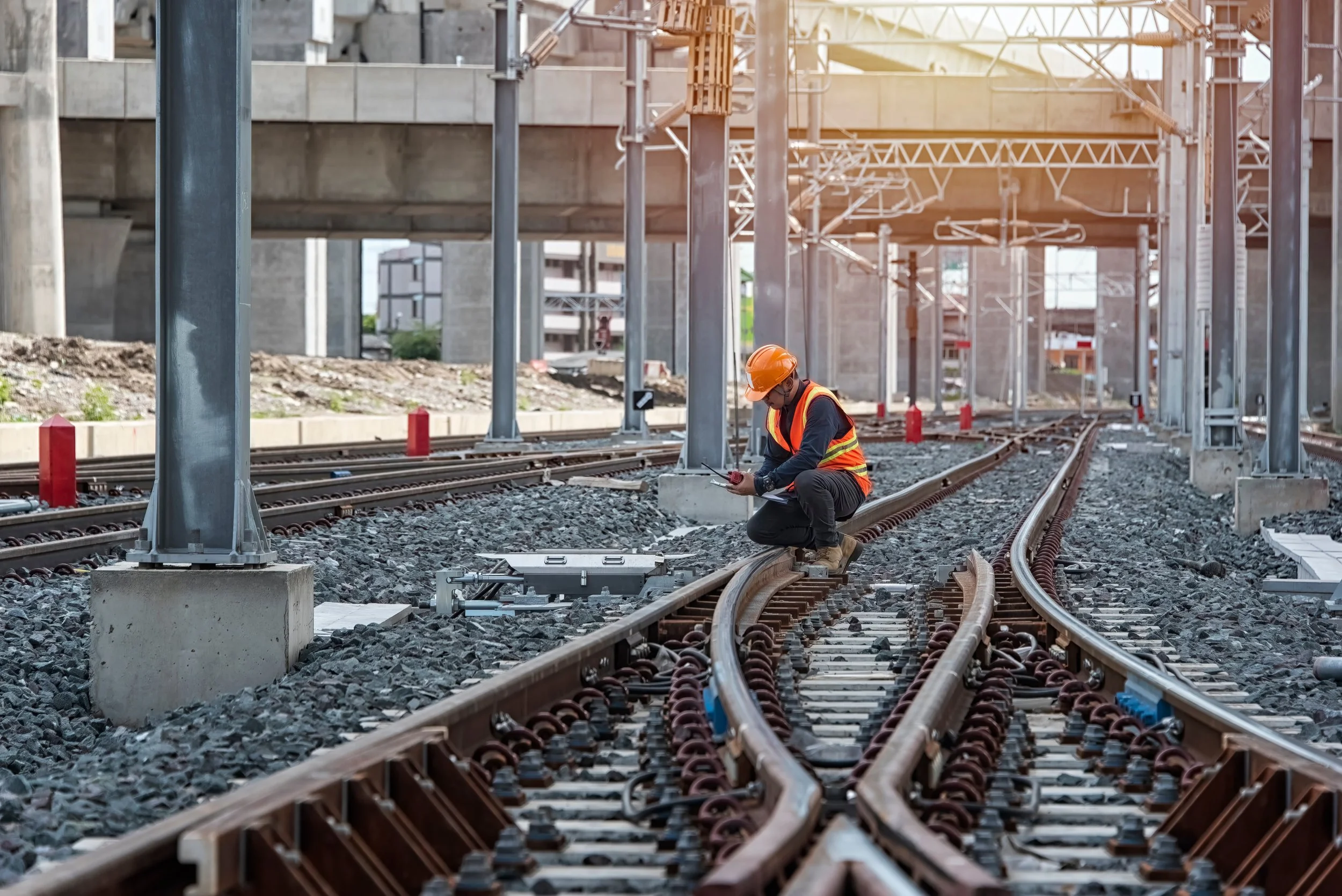 A construction worker wearing a safety helmet and vest kneels on railroad tracks at a construction site, inspecting or working on the tracks.