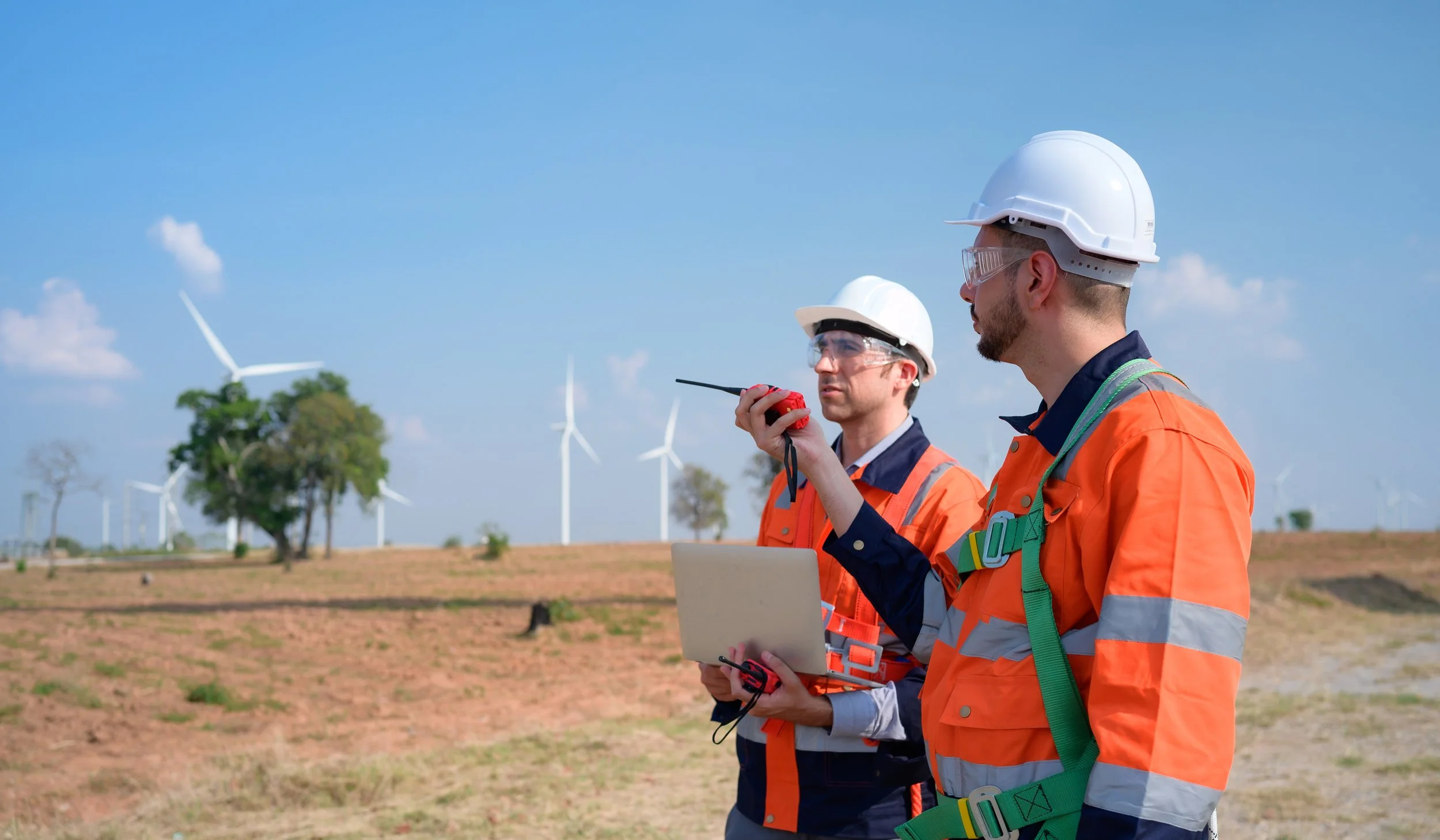 Two wind farm engineers inspecting wind turbines on a sunny day, wearing orange safety vests, white helmets, safety glasses, and holding a walkie-talkie and laptop.