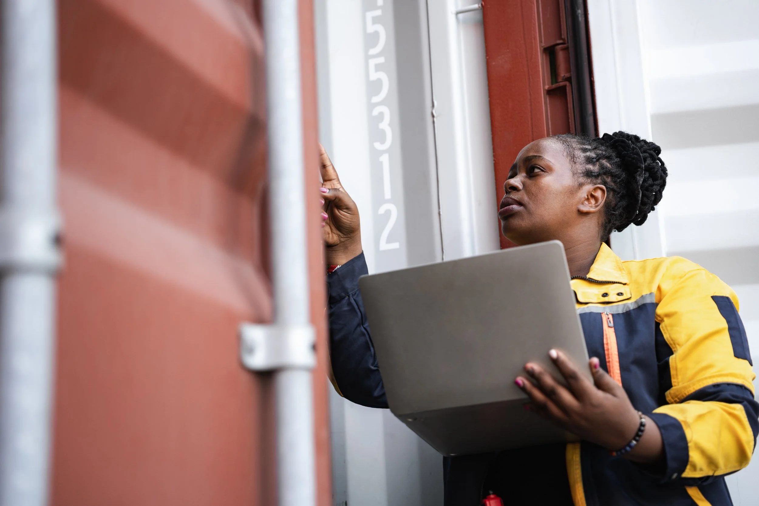 A woman with dark, curly hair in a bun, wearing a yellow and navy jacket, holding a gray laptop, and working on a delivery box on a wall.