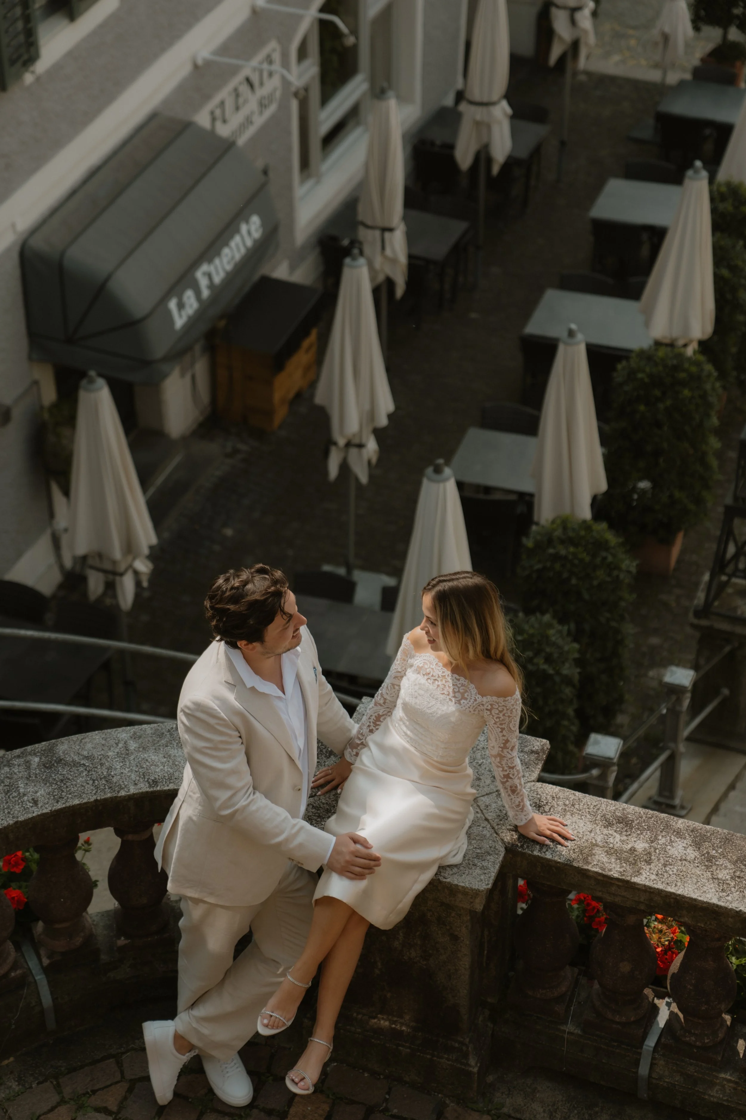 Ein Mann in einem weißen Anzug und eine Frau in einem weißen Hochzeitskleid sitzen auf einer Steinbrüstung auf einer Außenterrasse eines Restaurants namens "La Fontane". Sie unterhalten sich liebevoll, umgeben von weißen Schirmstationen und dekorativ