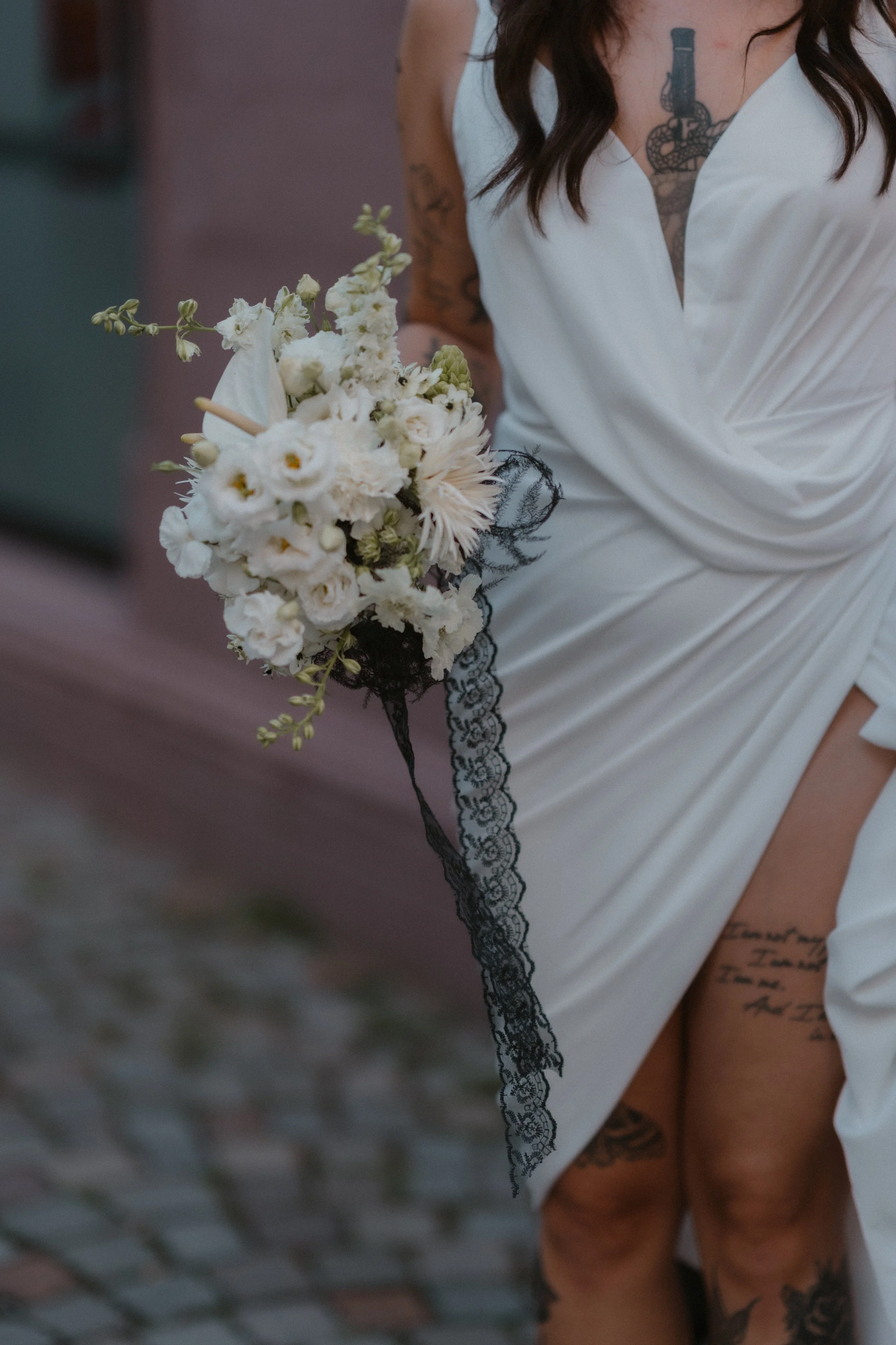 Nahaufnahme einer Frau in weißem Kleid, die einen weißen Blumenstrauß hält, mit sichtbaren Tattoos an Arm, Brust und Bein, und dunkelbraunem lockigem Haar.