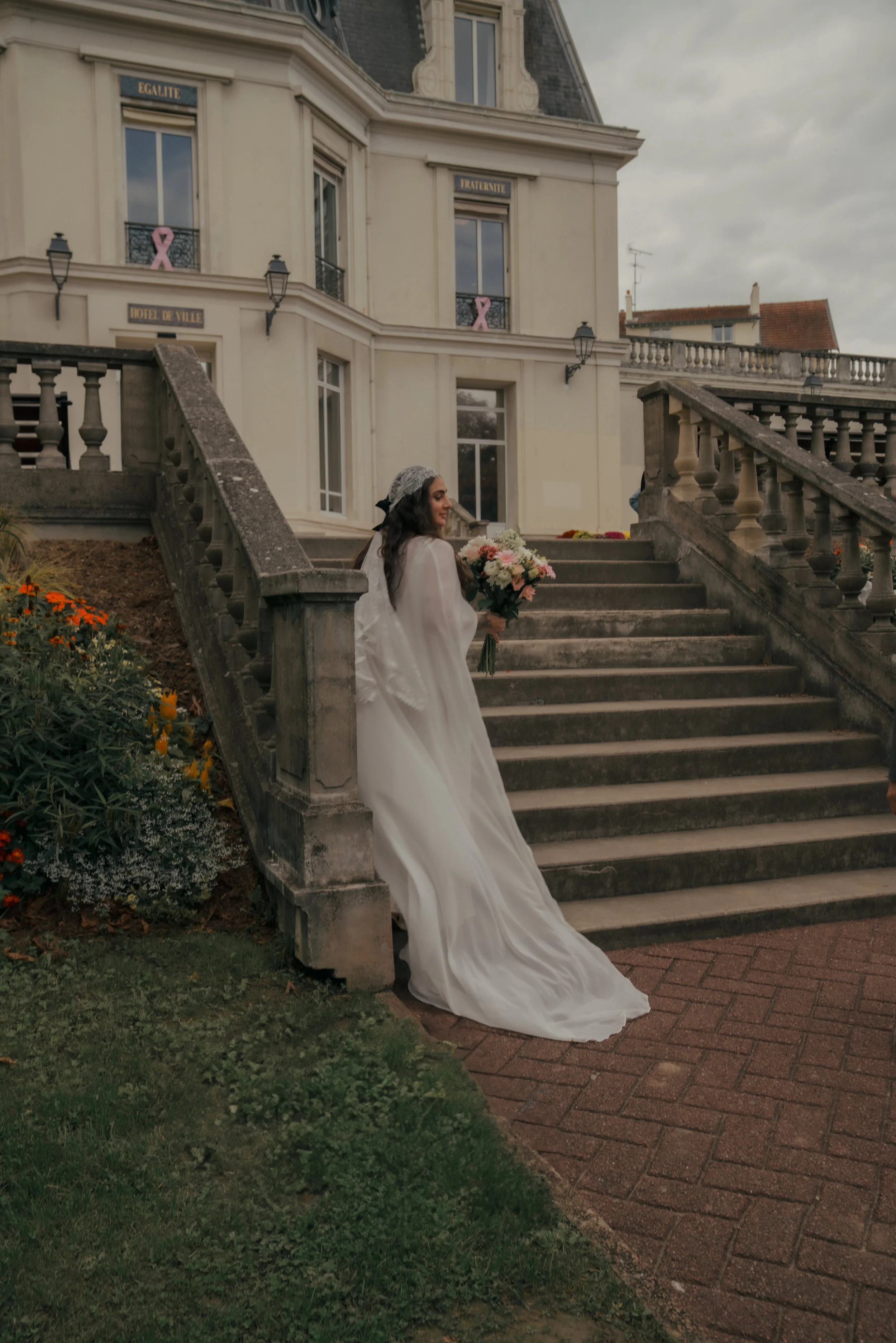 Eine Braut in einem weißen Kleid mit Blumenstrauß, die auf den Stufen eines stilvollen, weißen Gebäudes mit Balustraden steht.