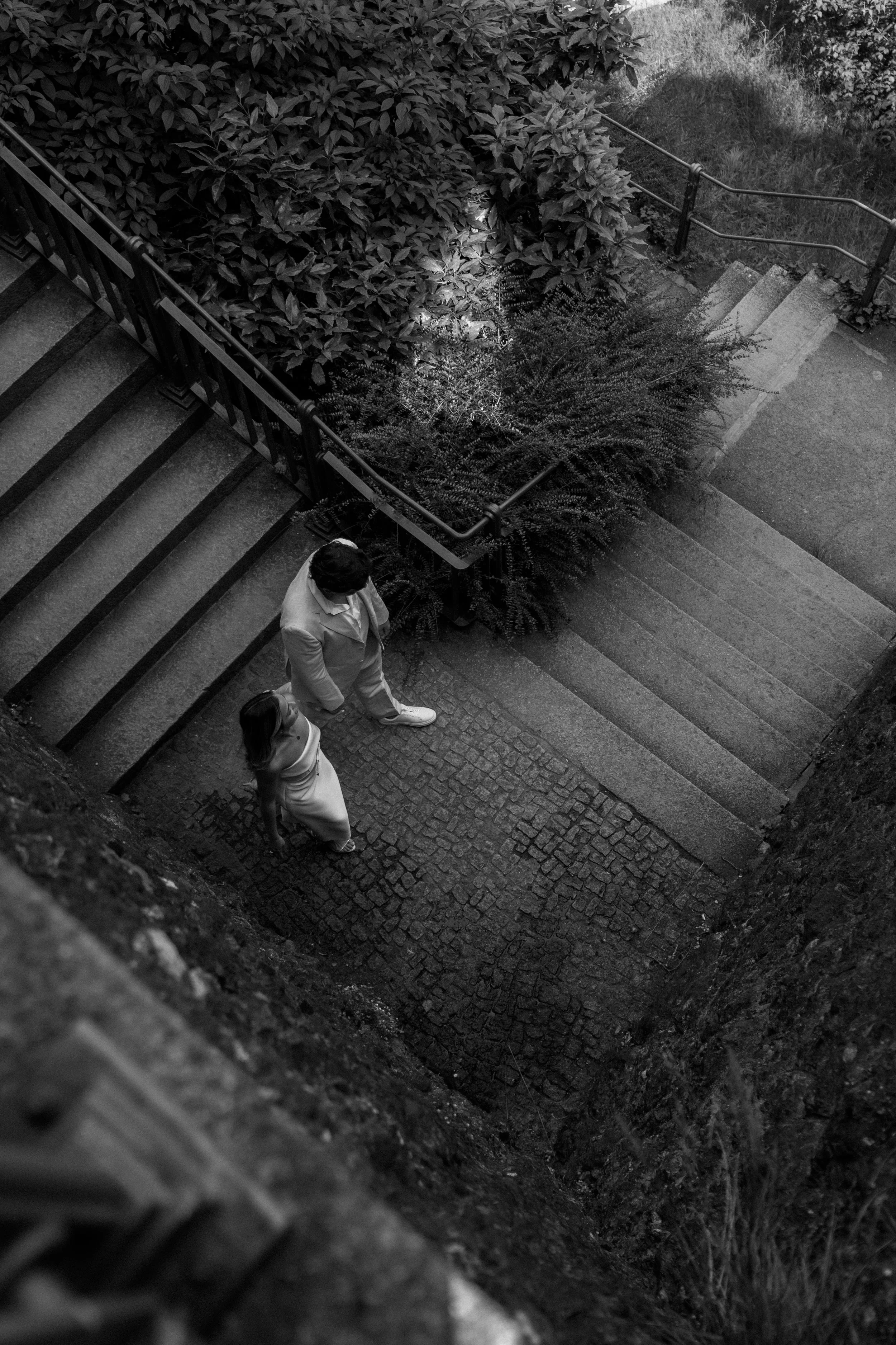 Zwei Menschen, ein Mann und eine Frau, stehen im Freien auf einer Treppe aus Stein, umgeben von viel Pflanzen und Büschen, in einer Schwarz-Weiß-Aufnahme.