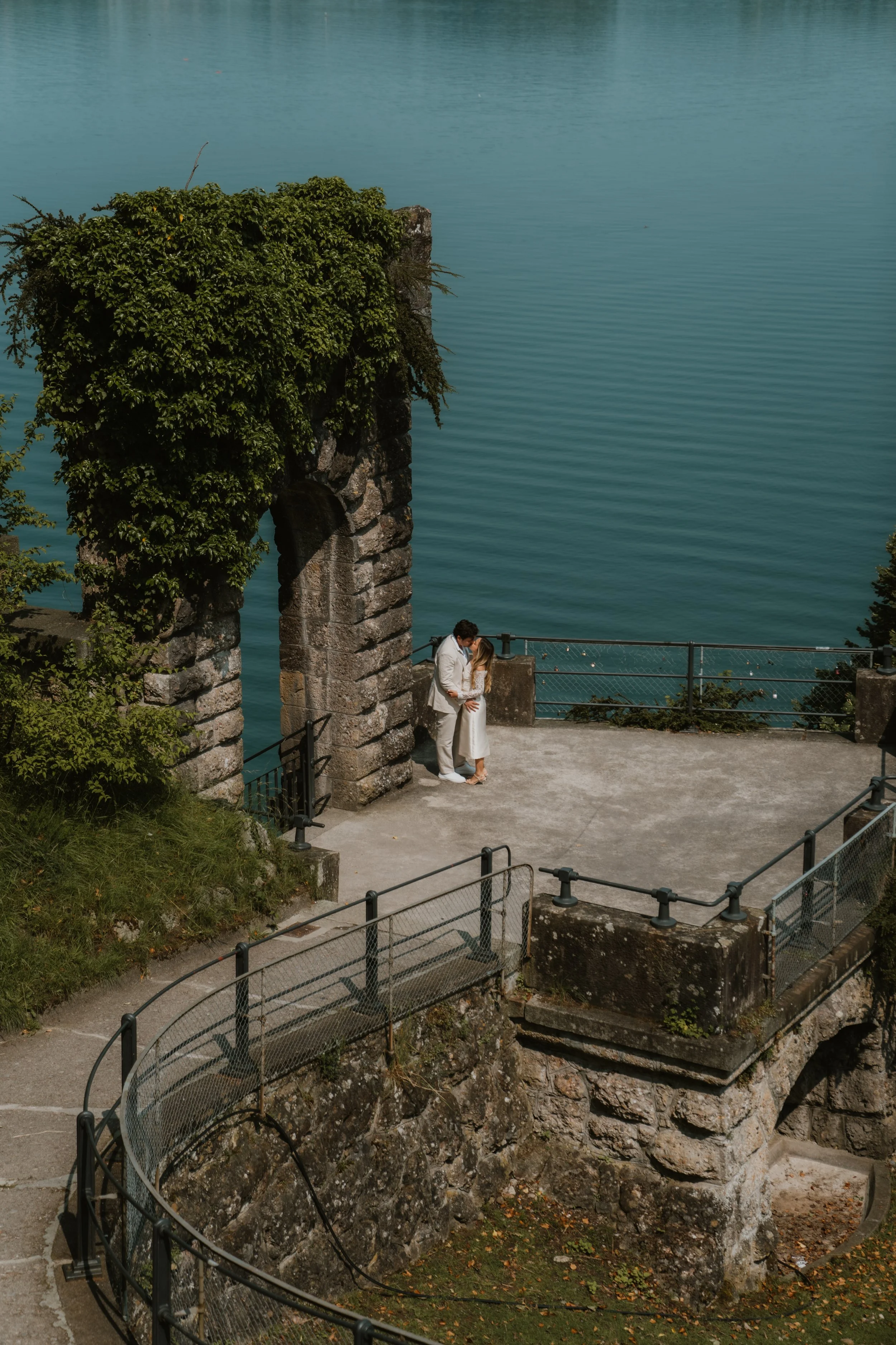 Ein verliebtes Paar umarmt sich vor einer alten Steinbrücke mit Blick auf einen ruhigen See, umgeben von Natur.