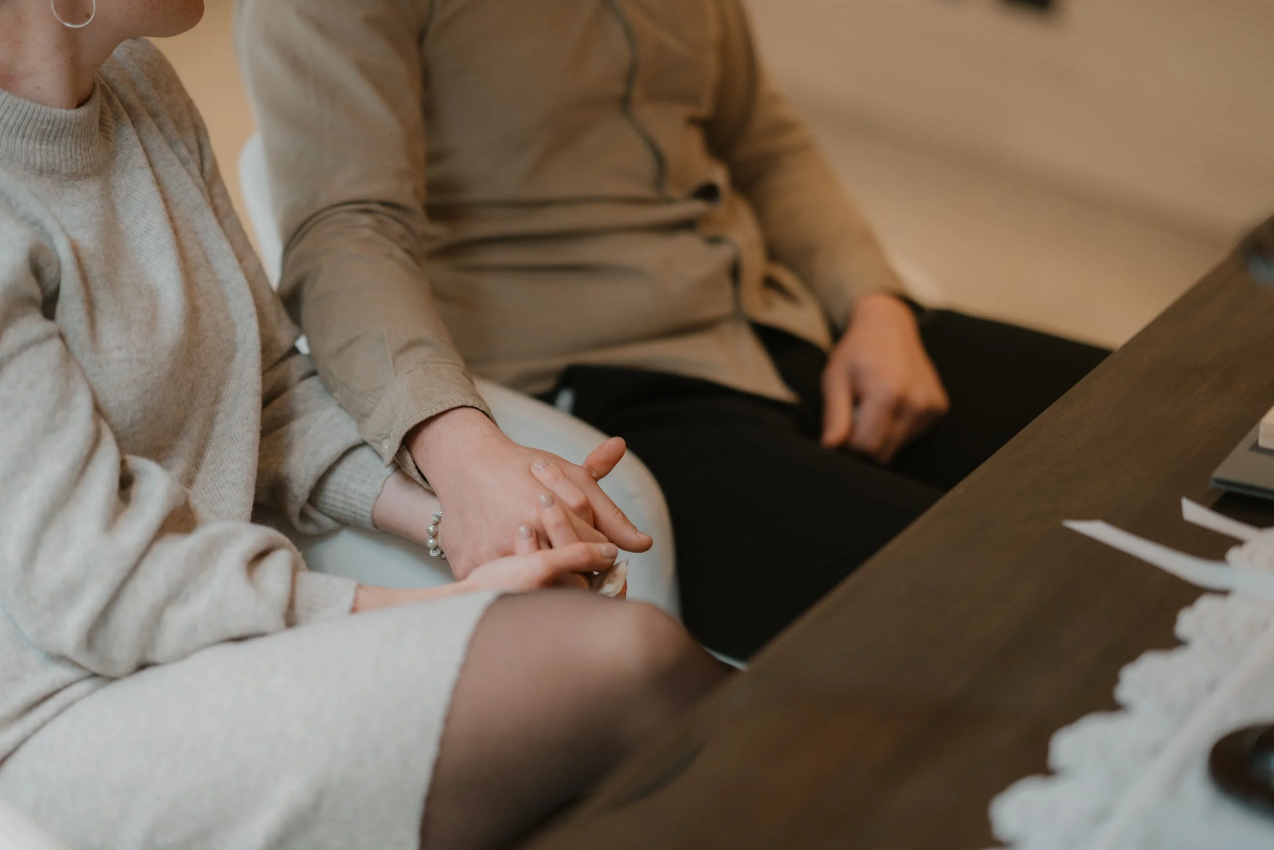 Zwei Menschen sitzen an einem Tisch und halten sich an den Händen. Im Vordergrund trägt eine Person eine beige Strickjacke, im Hintergrund eine beige Jacke.