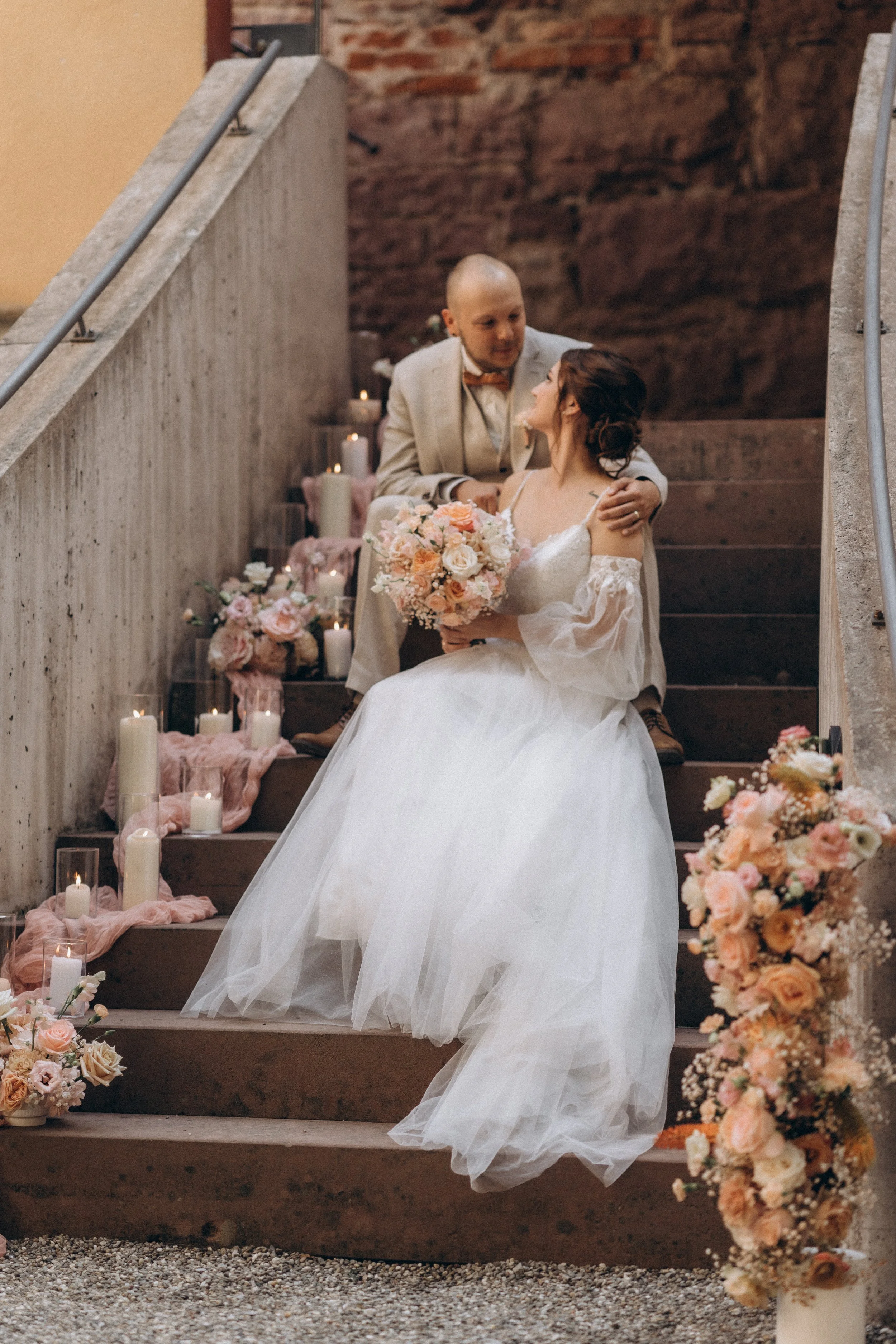 Ein Brautpaar sitzt auf einer Treppe, die mit Blumen und Kerzen dekoriert ist. Die Braut trägt ein weißes Hochzeitskleid, hält einen Blumenstrauß und schaut ihren Partner an. Der Bräutigam trägt einen hellen Anzug und schaut liebevoll die Braut an, w