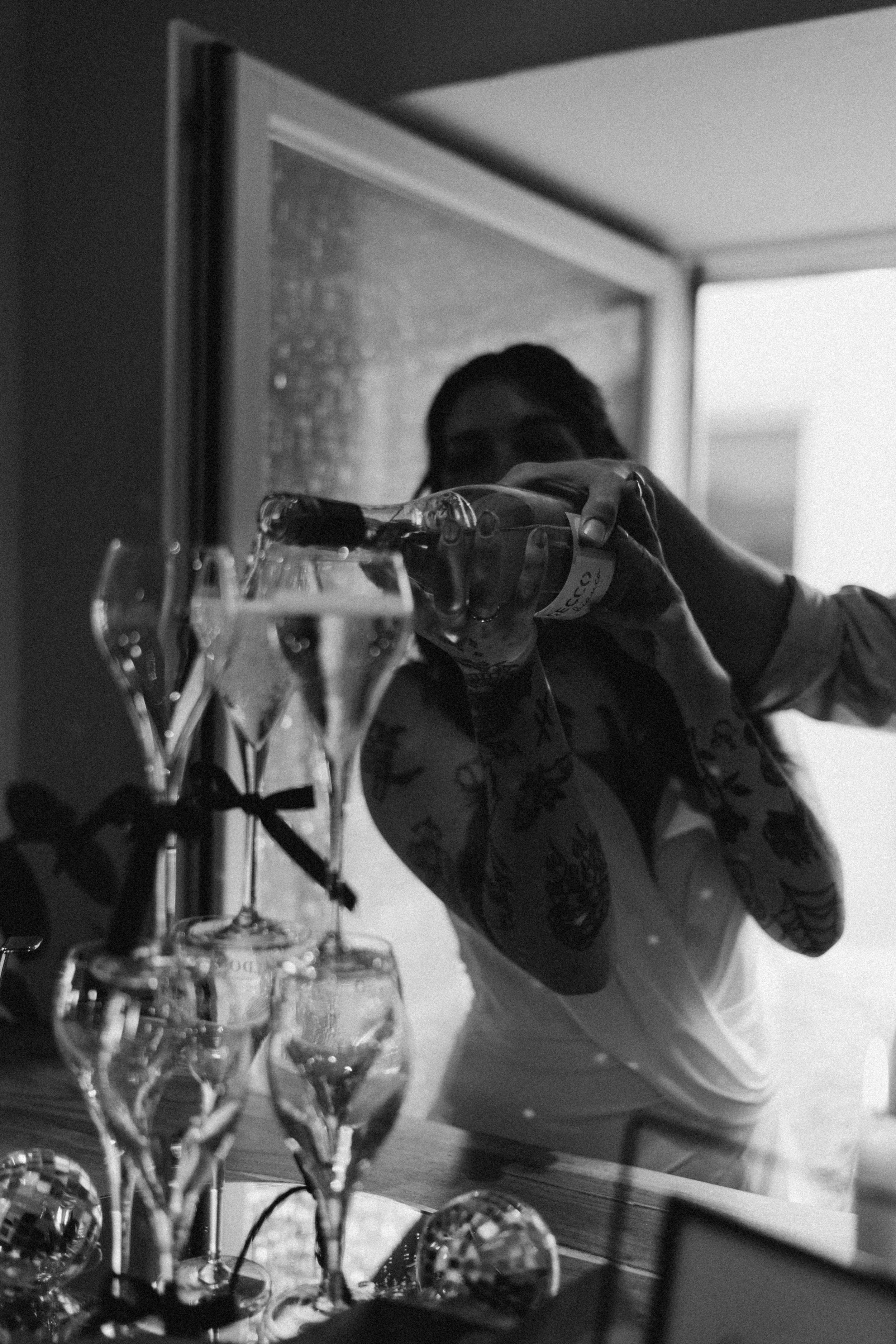 Schwarz-Weiß-Foto einer Person, die eine Champagnerflasche einschenkt, während sie durch ein Fenster blickt, mit mehreren leeren Sektgläsern auf einem Tisch im Vordergrund.