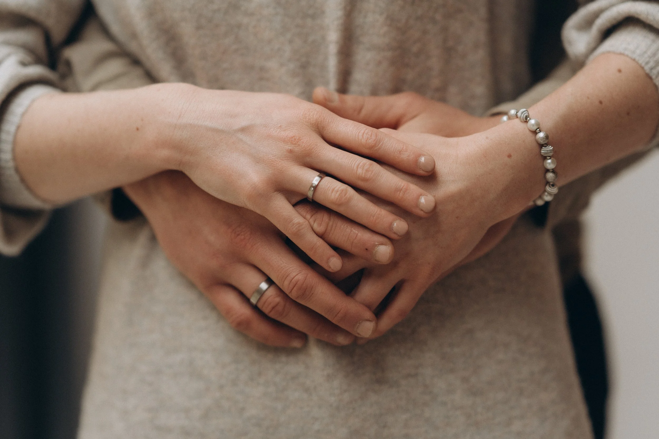 Nahaufnahme von zwei Händen, die sich berühren, mit Ringen an den Fingern und einer Perlenarmbanduhr am Handgelenk, vor einem hellen Stoff. संबंध herstellen.