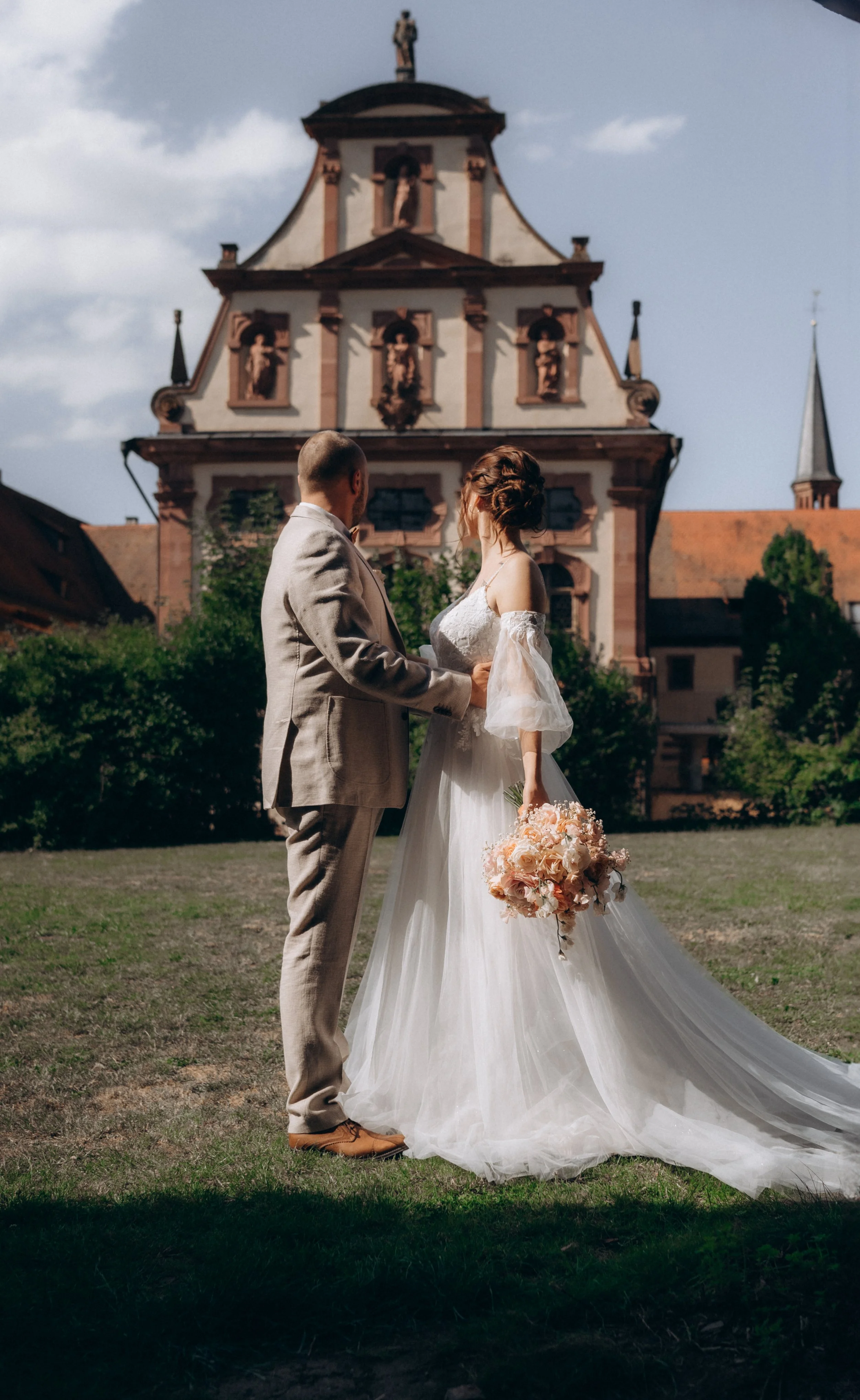 Braut und Bräutigam stehen sich vor einer historischen Kirche bei einer Hochzeit. Die Braut trägt ein weißes Brautkleid und hält einen Blumenstrauß, der Bräutigam trägt ein helles Sakko und braune Schuhe.