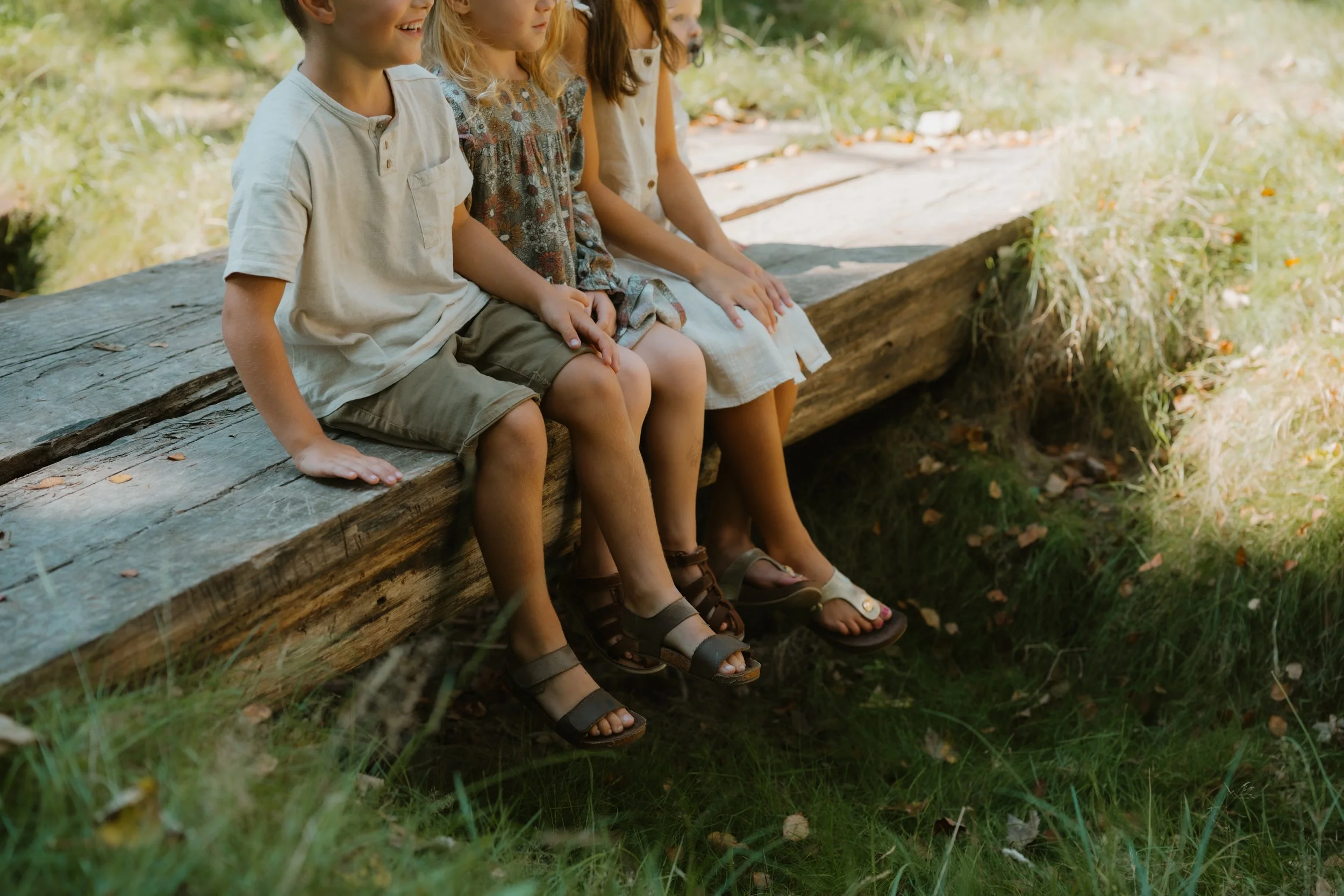 Vier Kinder sitzen nebeneinander auf einer Baumstamm im Grün, nur die unteren Hälften ihrer Körper sind sichtbar.