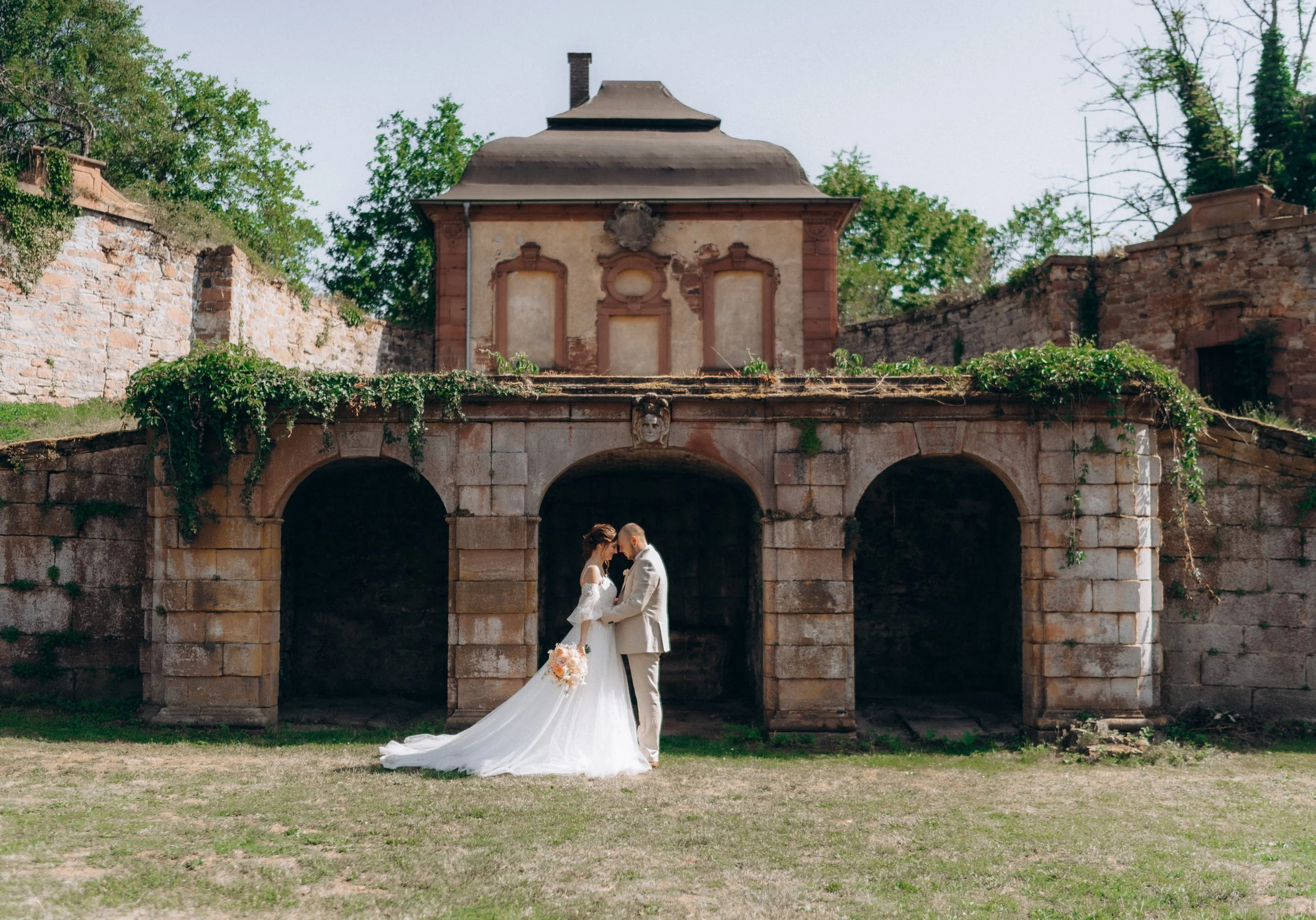 Ein Brautpaar steht sich unter einem alten, historischen Gebäude drückend, um die Hochzeit zu feiern. Das Gebäude hat einen Balkon mit Bogen und ist umgeben von grünen Bäumen.