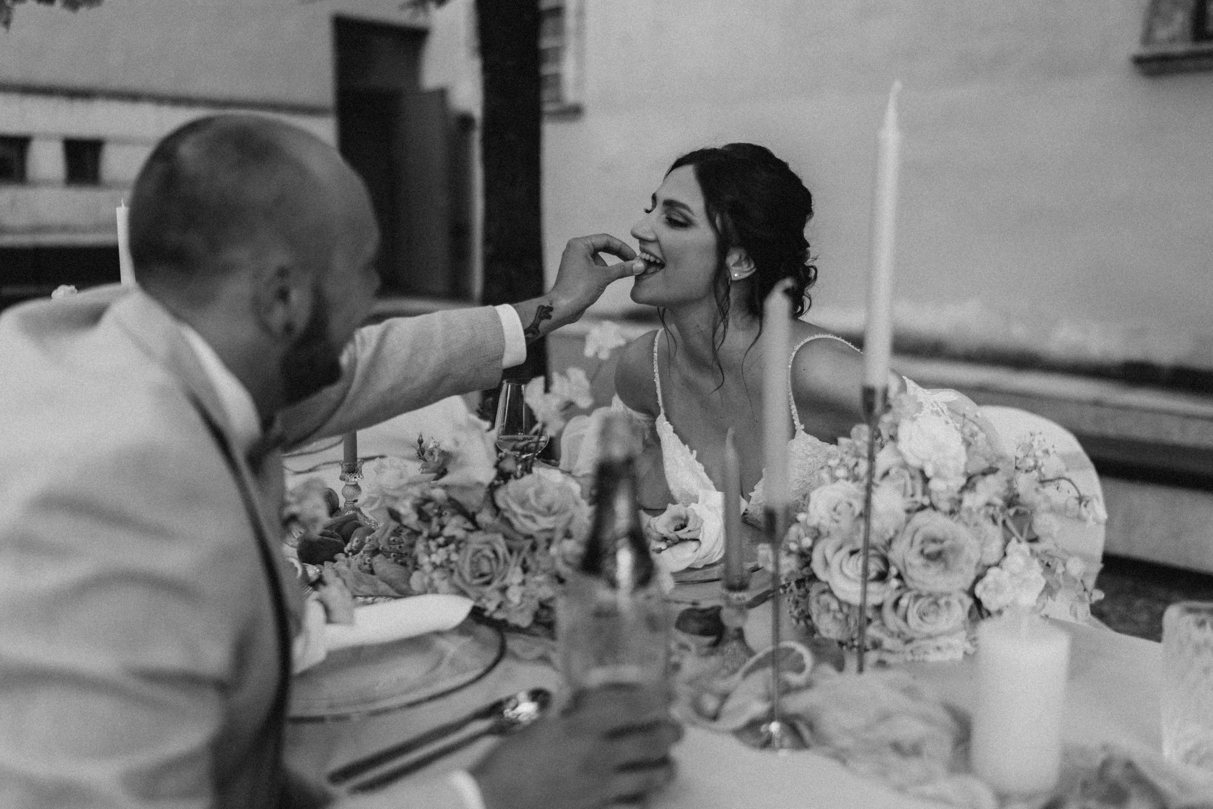 Schwarz-weiß-Bild von einem Hochzeitsessen, bei dem ein Brautpaar sitzt. Eine Frau in einem Hochzeitskleid lächelt, während eine andere Person ihr einen Kuss auf die Wange gibt. Es gibt Kerzen und Blumen auf dem Tisch.