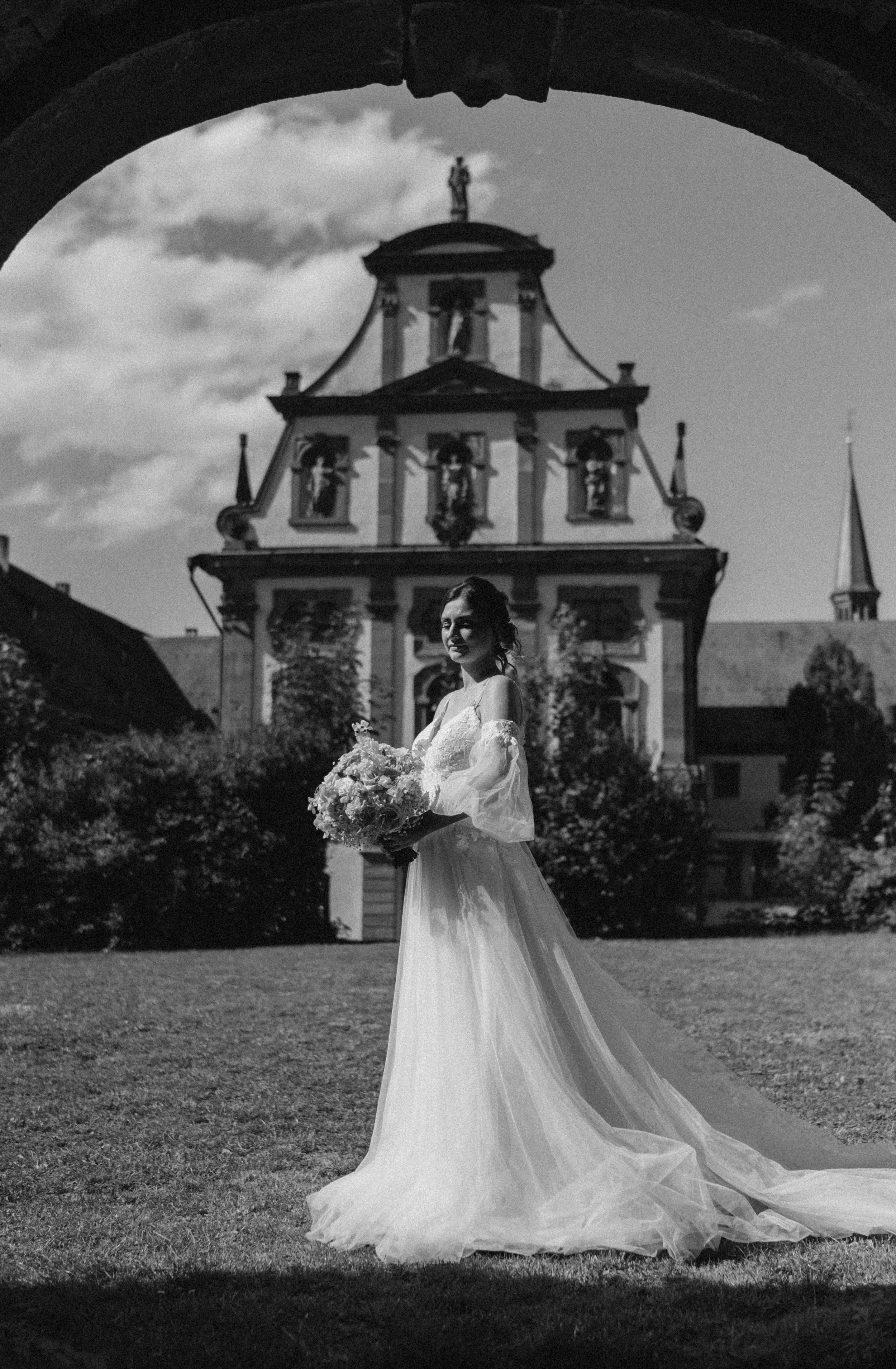 Schwarz-Weiß-Foto einer Frau im Brautkleid mit Brautstrauß vor einer historischen Kulisse.
