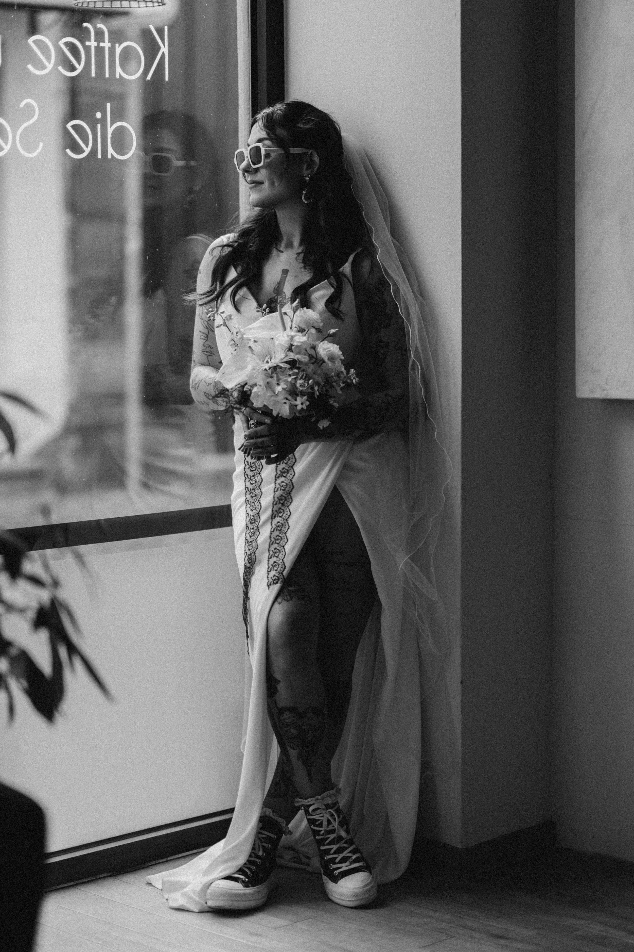 Schwarz-weiß-Foto einer Frau im Hochzeitskleid, die mit Sonnenbrille und Tattoos auf einem Bürgersteig steht. Sie hält einen Blumenstrauß und trägt Sneakers. Im Hintergrund ist eine Glasscheibe mit weißen Schriftzug.