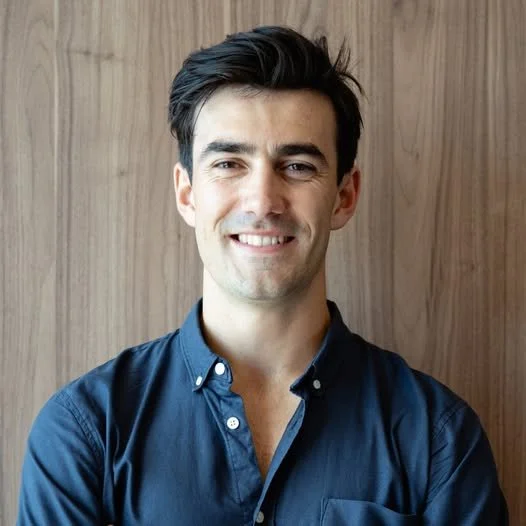 Reuben smiles and is wearing a dark blue collared shirt. He has black hair.