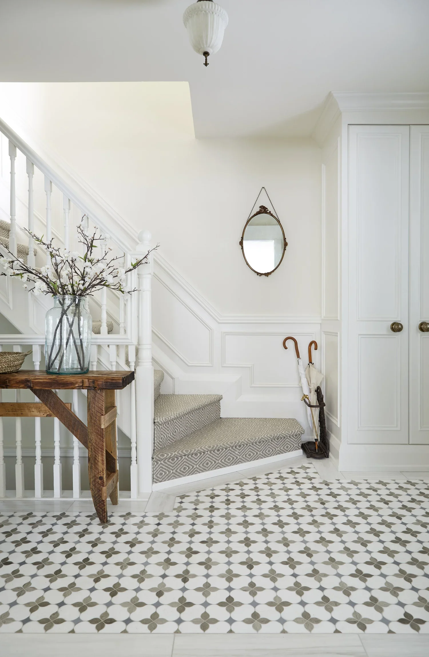 Entrance hallway with patterned tile floor, white walls, a staircase with a carpet runner, a wooden table with a glass vase of branches, a mirror hanging on the wall, and umbrellas near a cabinet.