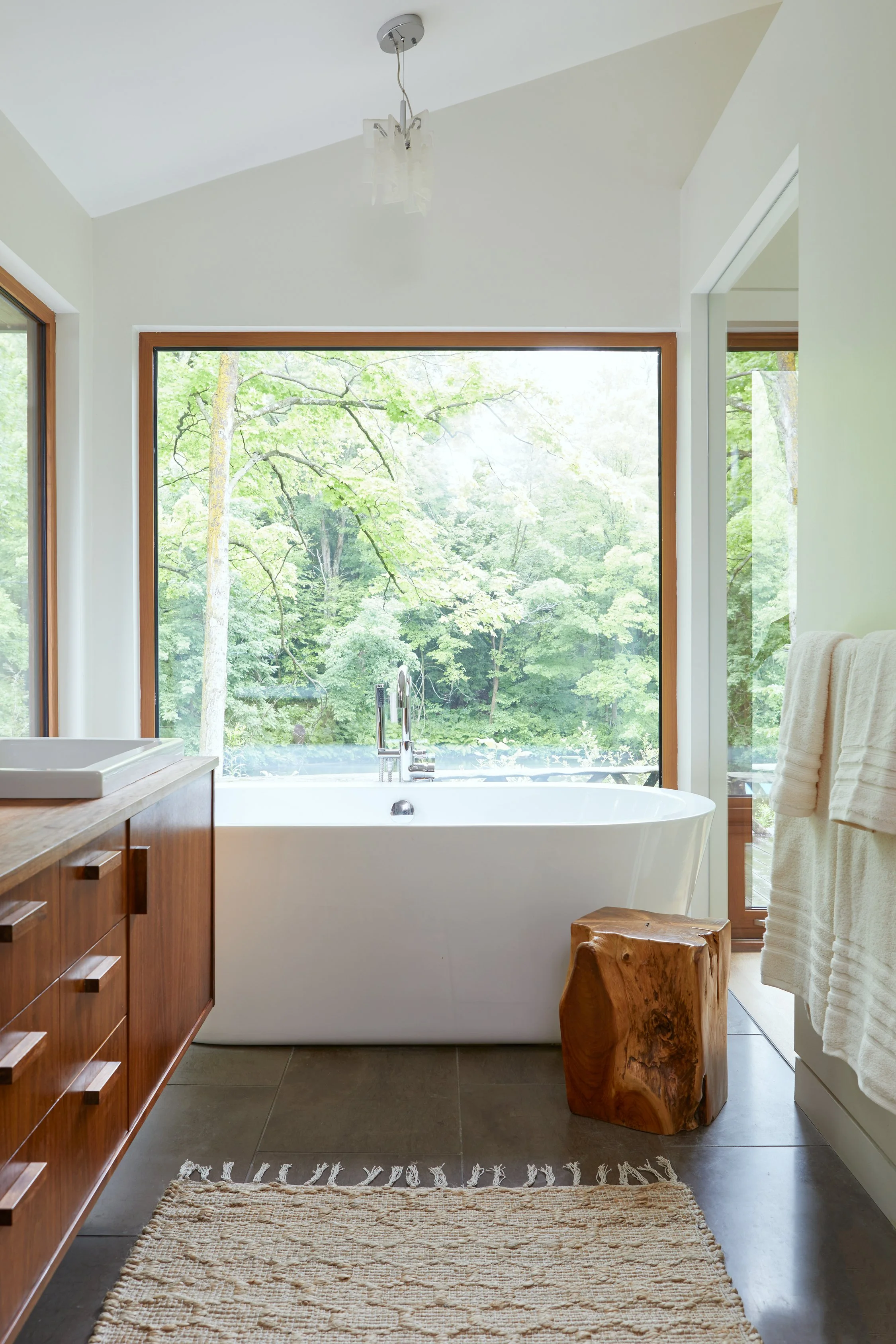 Bright bathroom with a large window showing green trees outside, a white bathtub, a wooden bathroom vanity, a wooden stool, white towels, and a beige rug.
