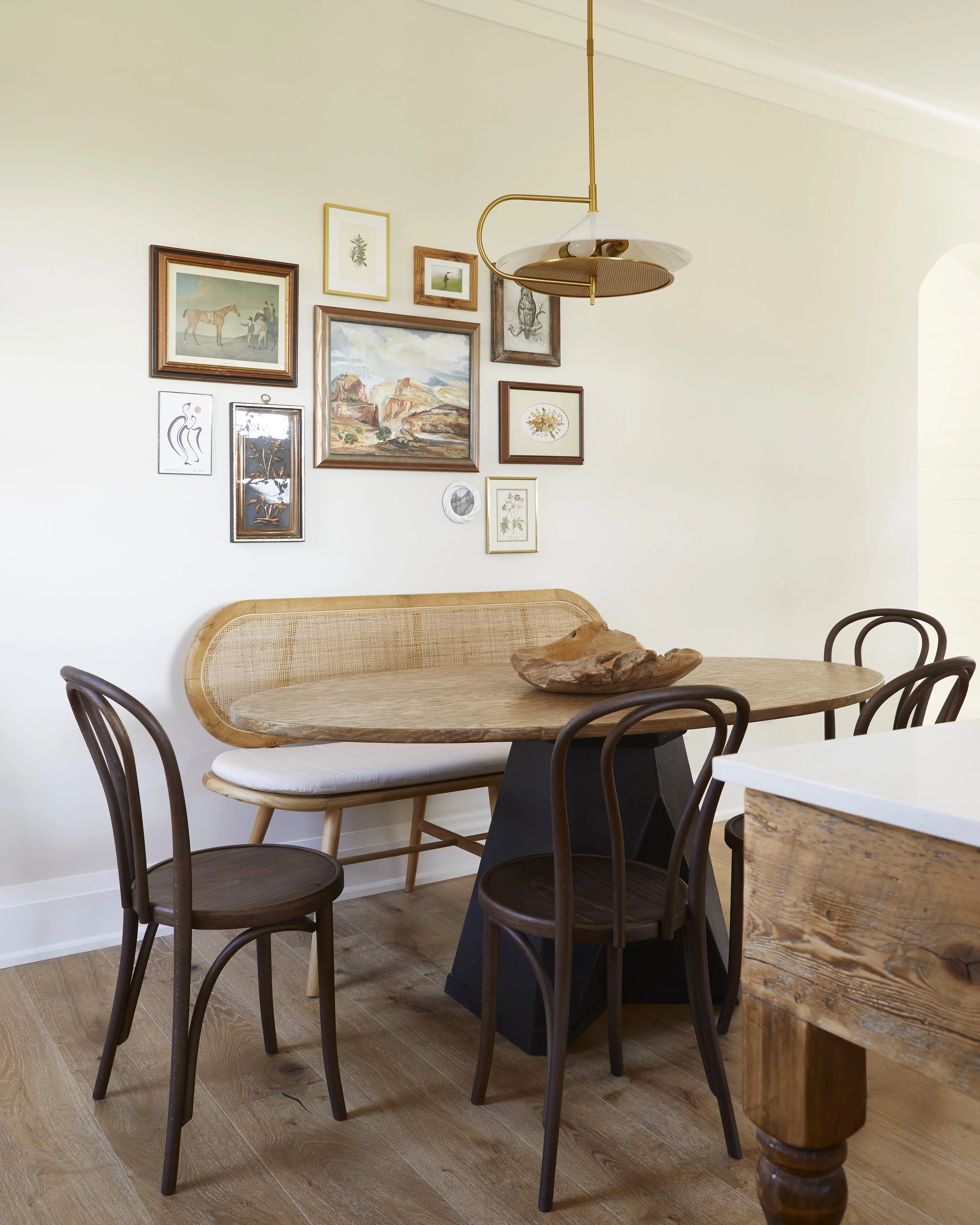 A dining area with a wooden table, four dark wooden chairs, a built-in wooden bench with a cushion, and art gallery wall with various framed pictures, pendant light fixture, and a wood bowl centerpiece.