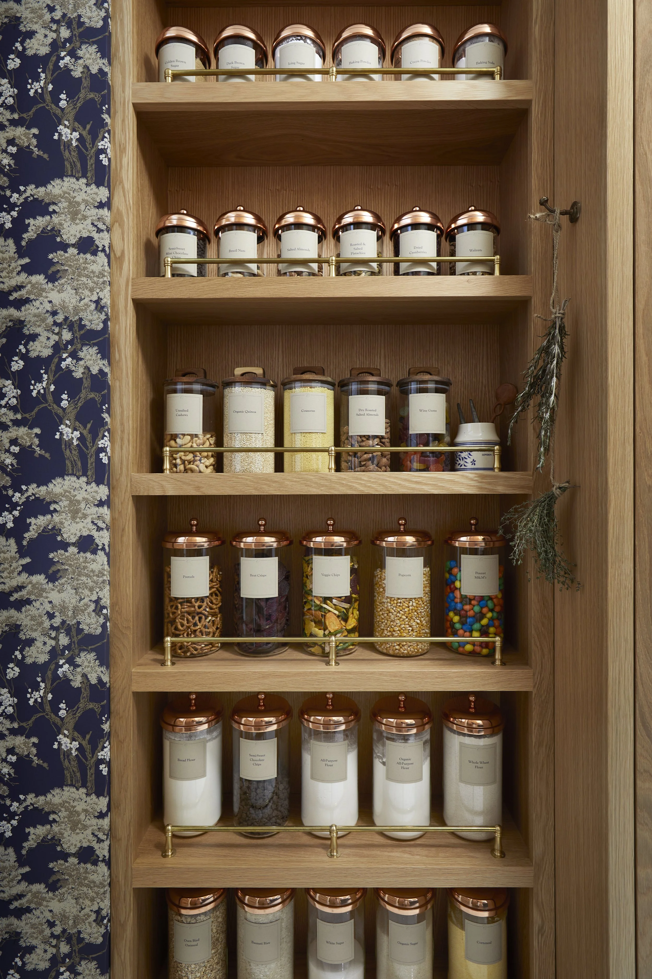 A wooden pantry cabinet with five shelves holding labeled glass jars filled with various baking ingredients and toppings like sugar, flour, nuts, sprinkles, and candies.