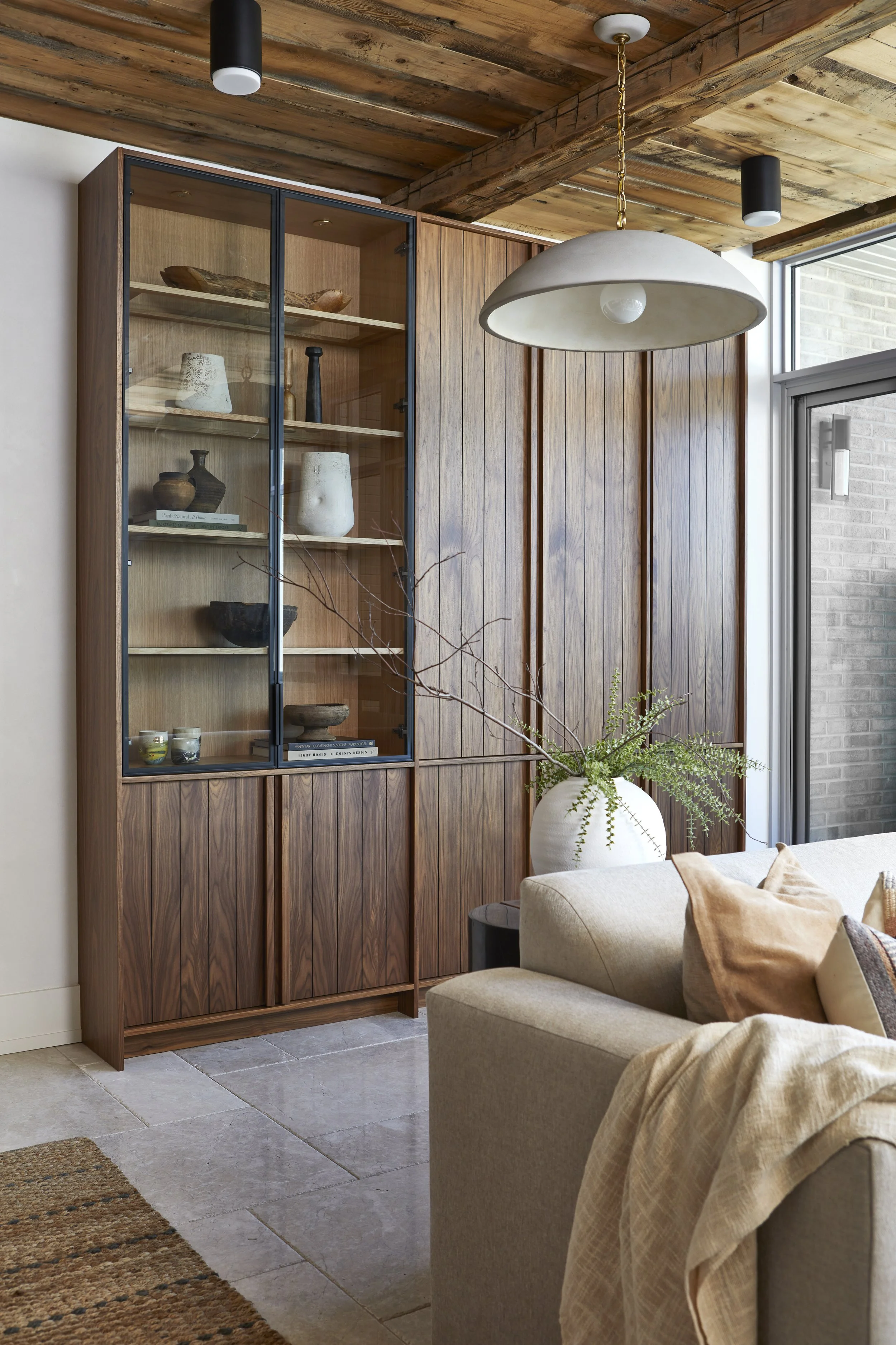 A living room interior featuring a wooden cabinet with glass doors displaying pottery and books, a beige sofa with throw pillows, a large white vase with greenery, a pendant ceiling light, and a wooden ceiling with exposed beams near a window with a 