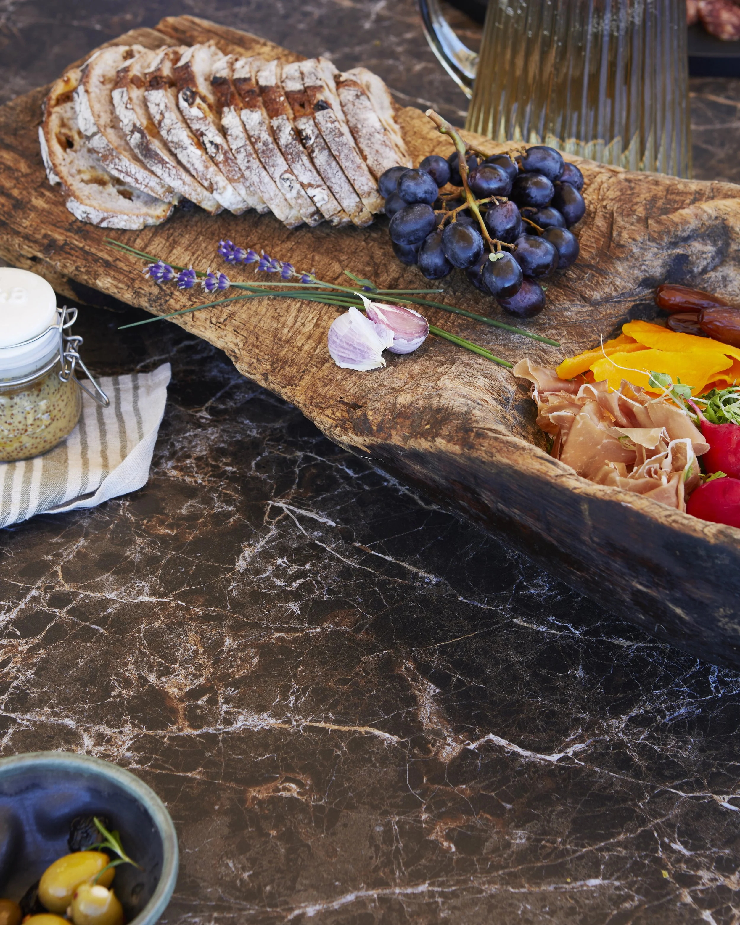 A wooden serving board with slices of fruitcake, a bunch of black grapes, garlic, lavender, and a cheese spread in a small jar. On the side, there is a bowl of olives.