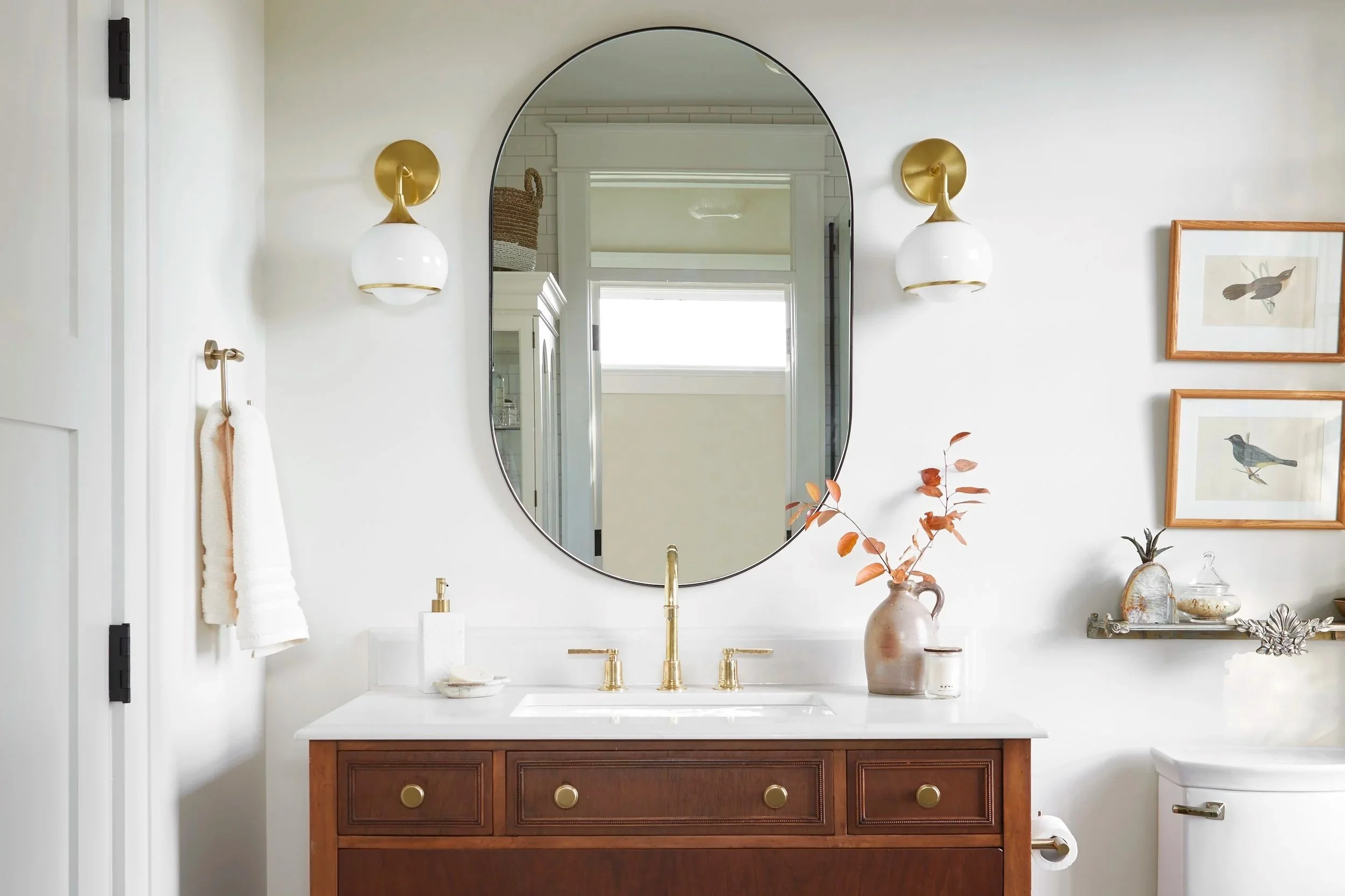 A bathroom featuring a wooden vanity with a white countertop, gold fixtures, an oval mirror, wall sconces, and decorative decor including framed bird artwork, a vase with dried leaves, and a shelf with various items.