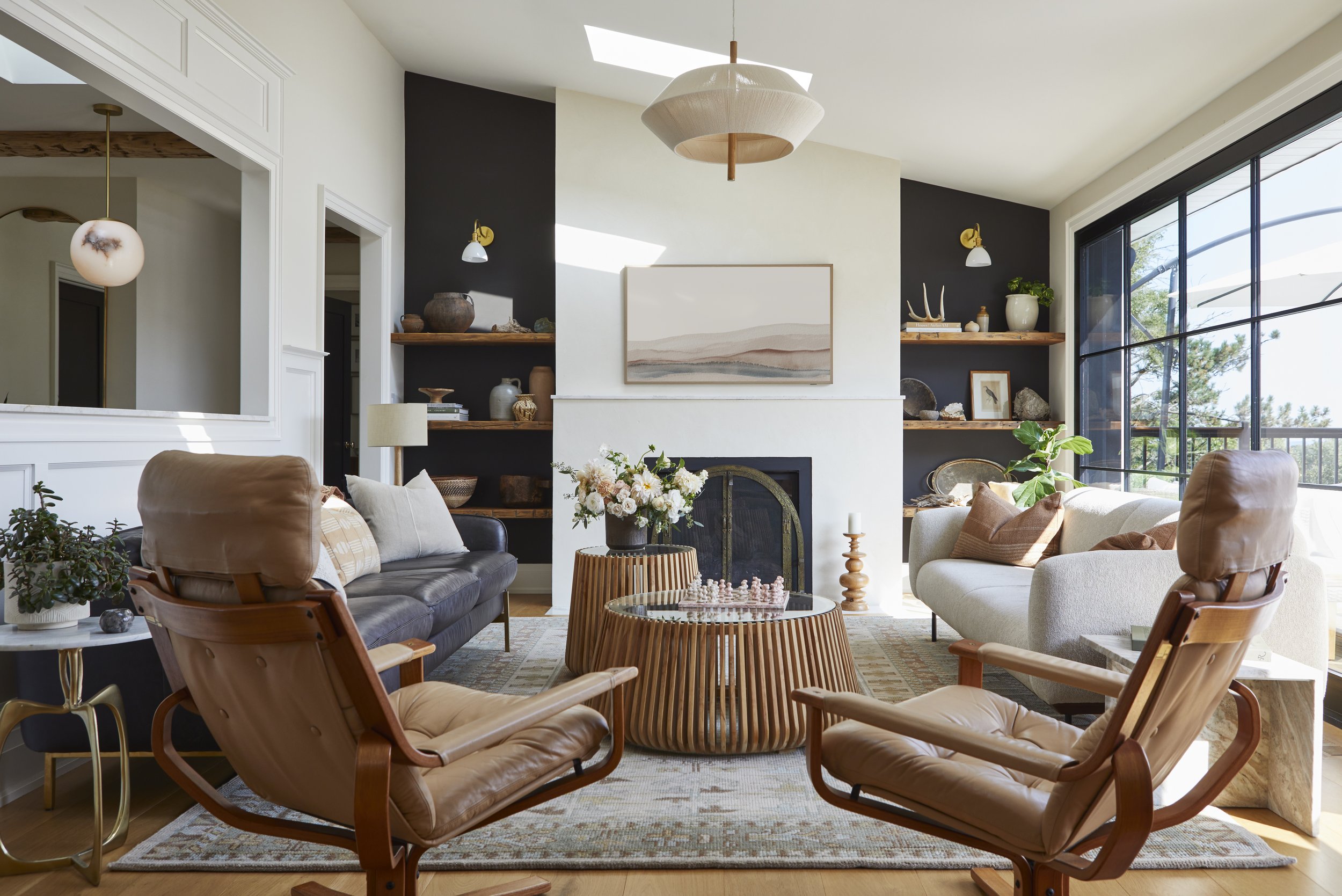 Living room with two leather armchairs, a black sofa, a white sofa, and a wooden coffee table with a chess set, fireplace, built-in shelves with decor, large window showing greenery outside, and ceiling light fixtures.
