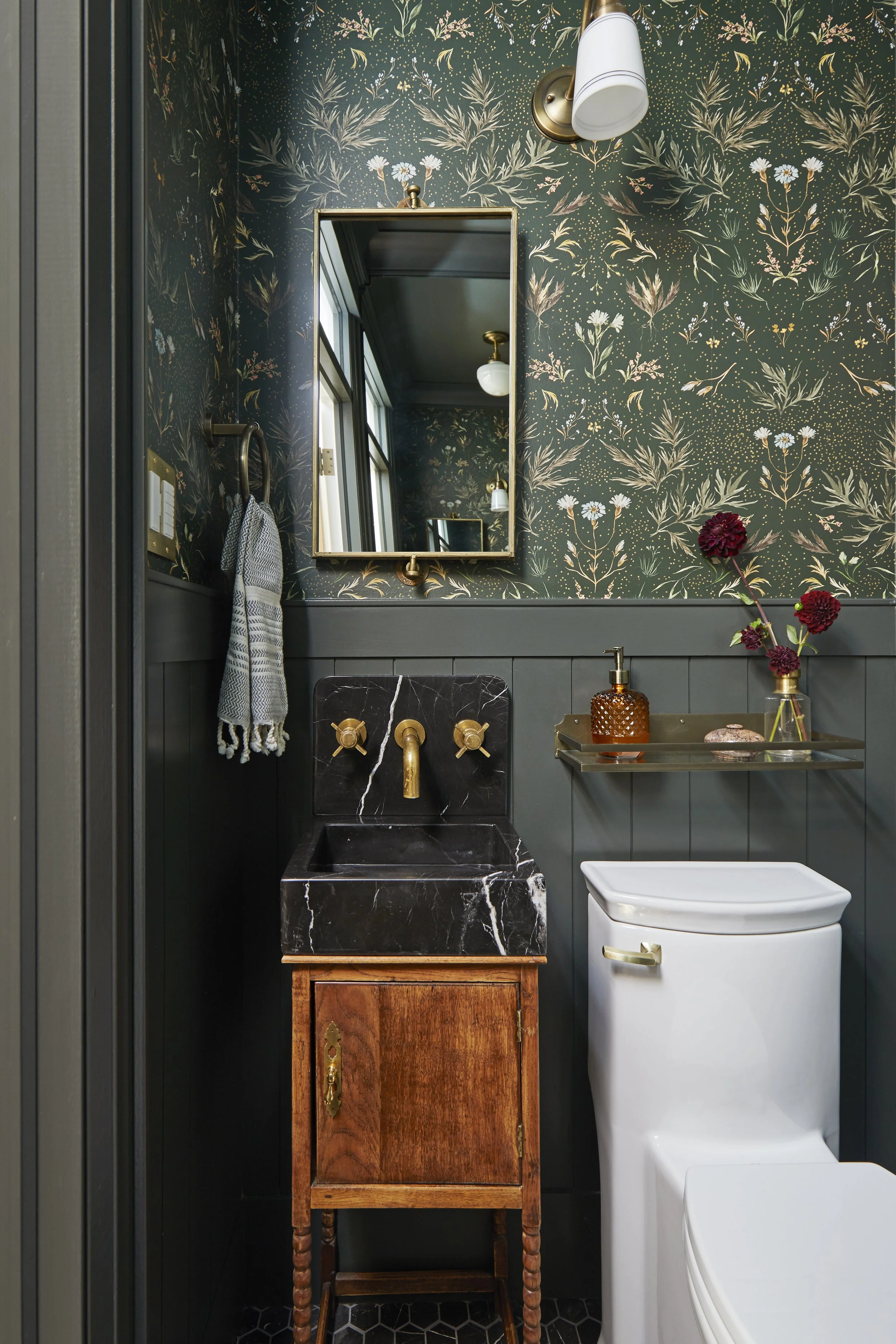 A small bathroom with dark floral wallpaper, a mirror, a brass wall light, a gray towel, a black marble sink on a wooden cabinet, a gold shelf with a amber soap dispenser, a vase with red flowers, and a white toilet.
