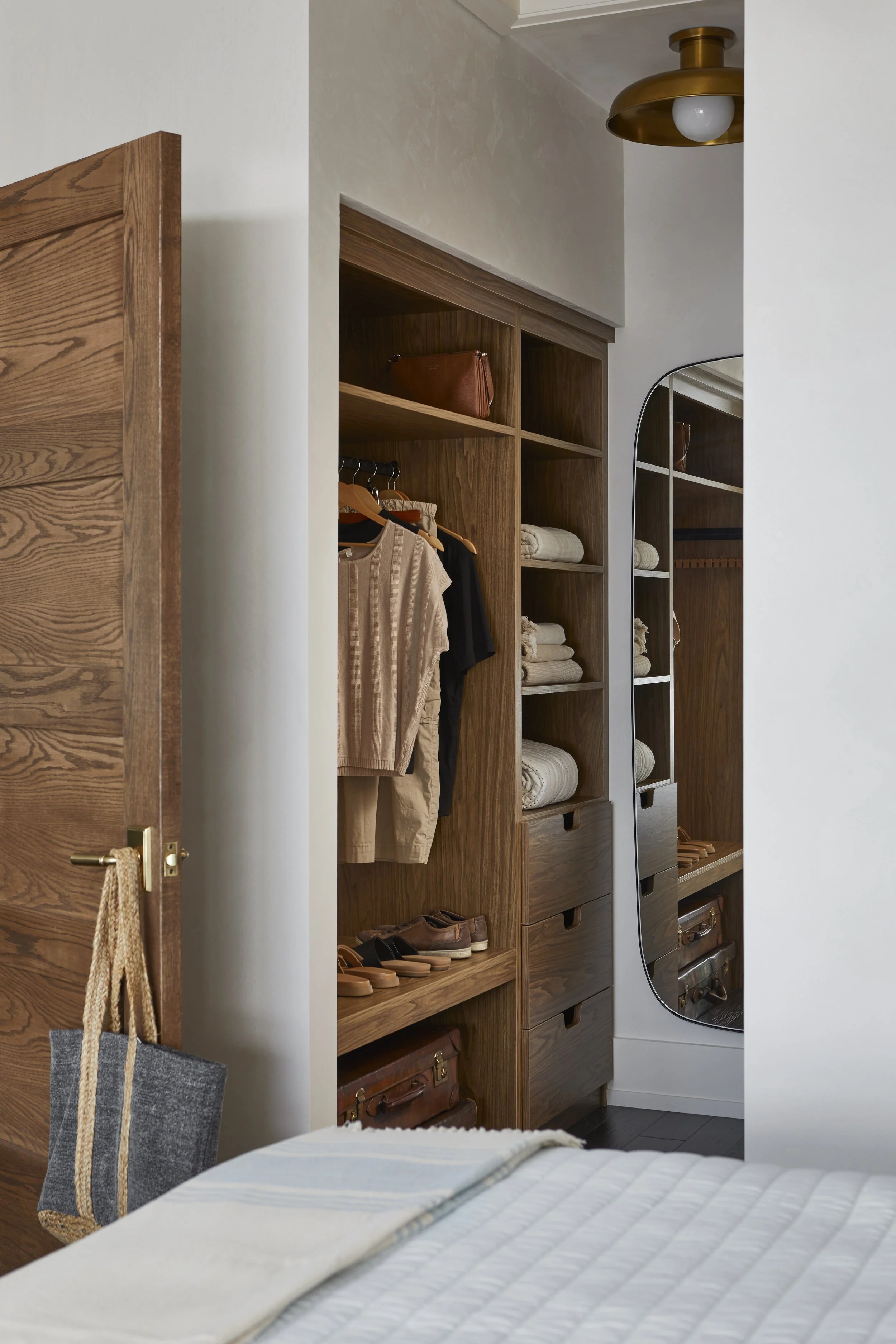 View of a closet with wooden shelves filled with folded towels, hanging clothes, shoes, and a leather bag, reflected in a full-length mirror.