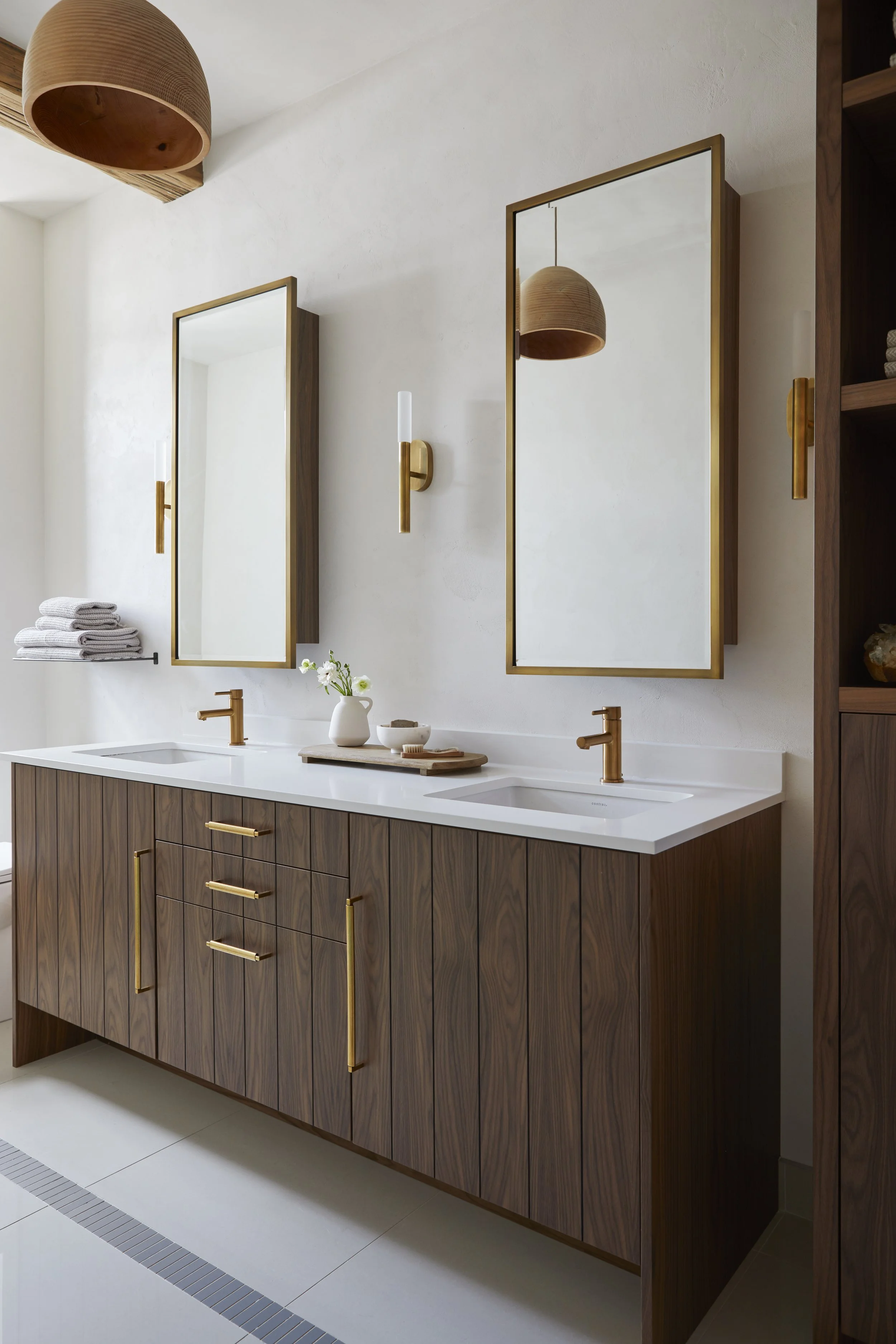 Modern bathroom with double vanity, wooden cabinets with gold handles, white countertop, two mirrors with gold frames, gold faucet fixtures, wall sconce lights, towels on a rack, decorative items on the counter, and a wooden pendant light