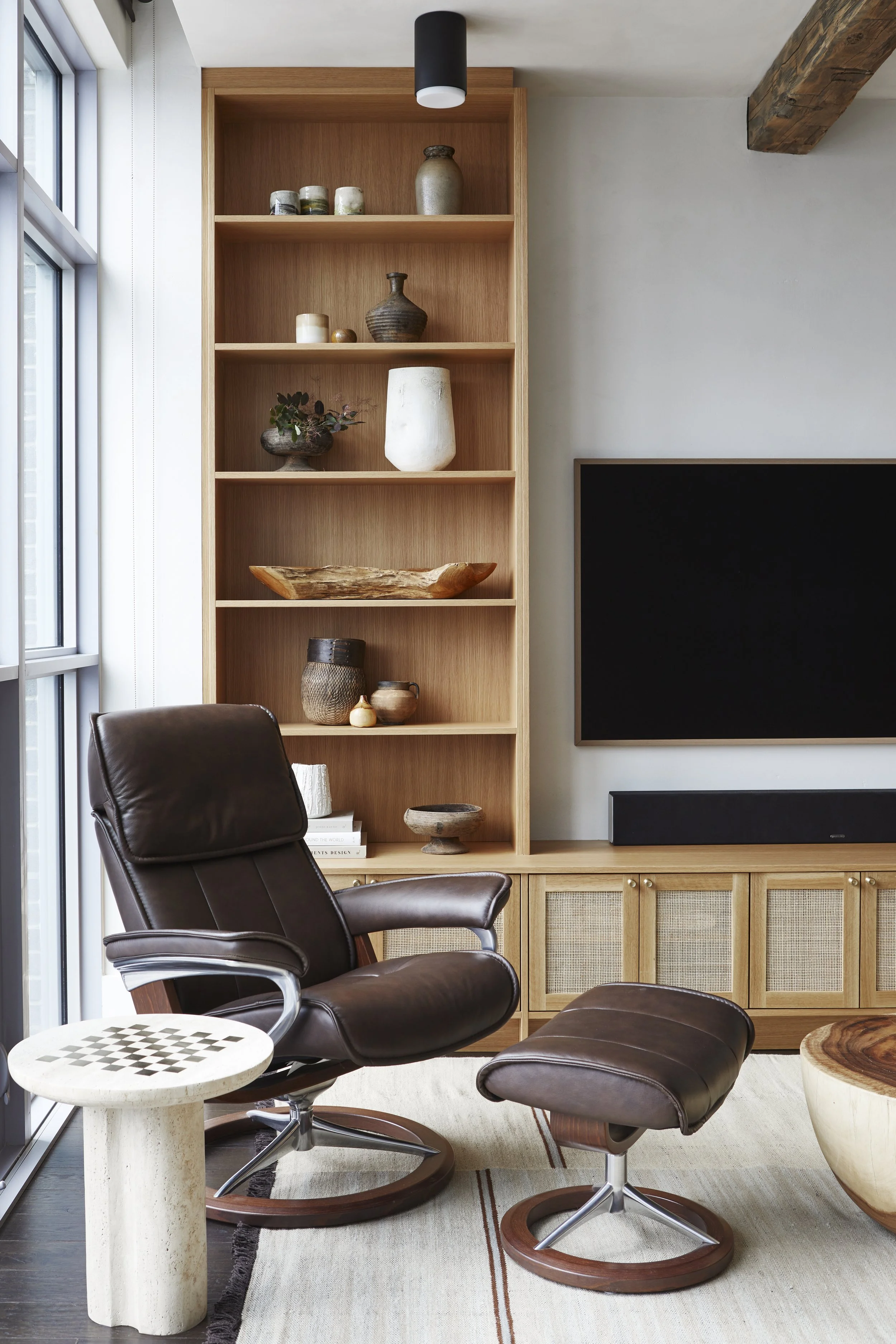 A living room with a brown leather swivel chair and matching ottoman, a cream-colored side table with a chessboard pattern, a wooden storage unit with woven panels, and a large black television. There is a built-in wooden bookshelf with decorative va