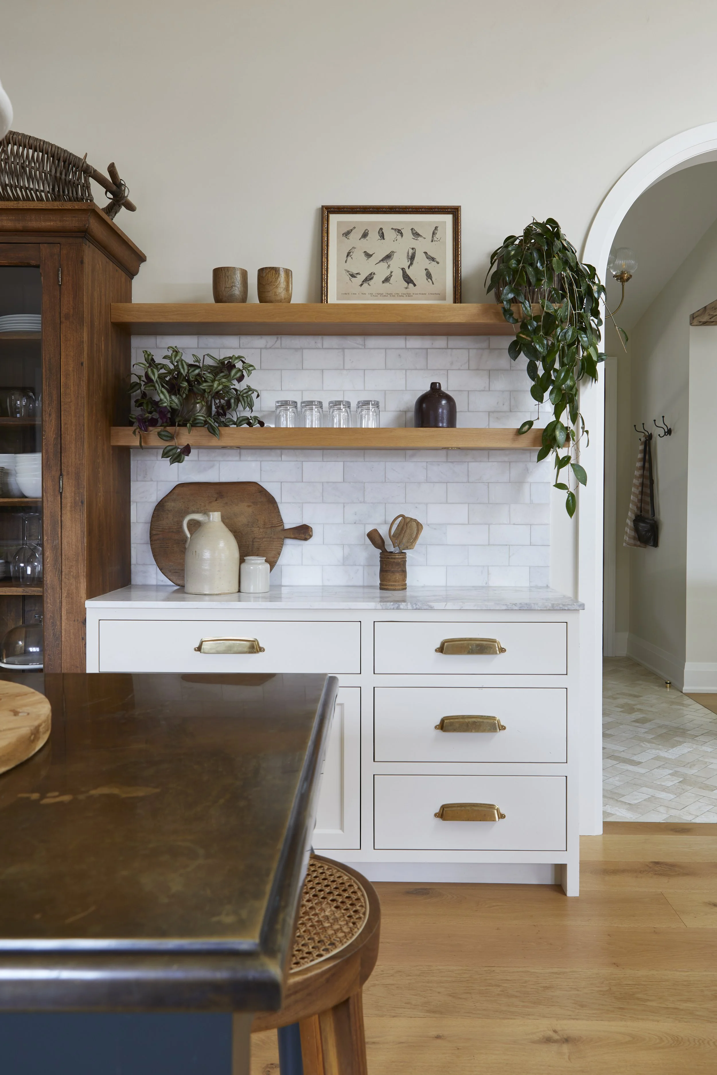 A kitchen with a white cabinet, wooden open shelves, and various decorative items including plants, pottery, and framed pictures, with a wooden table in the foreground.