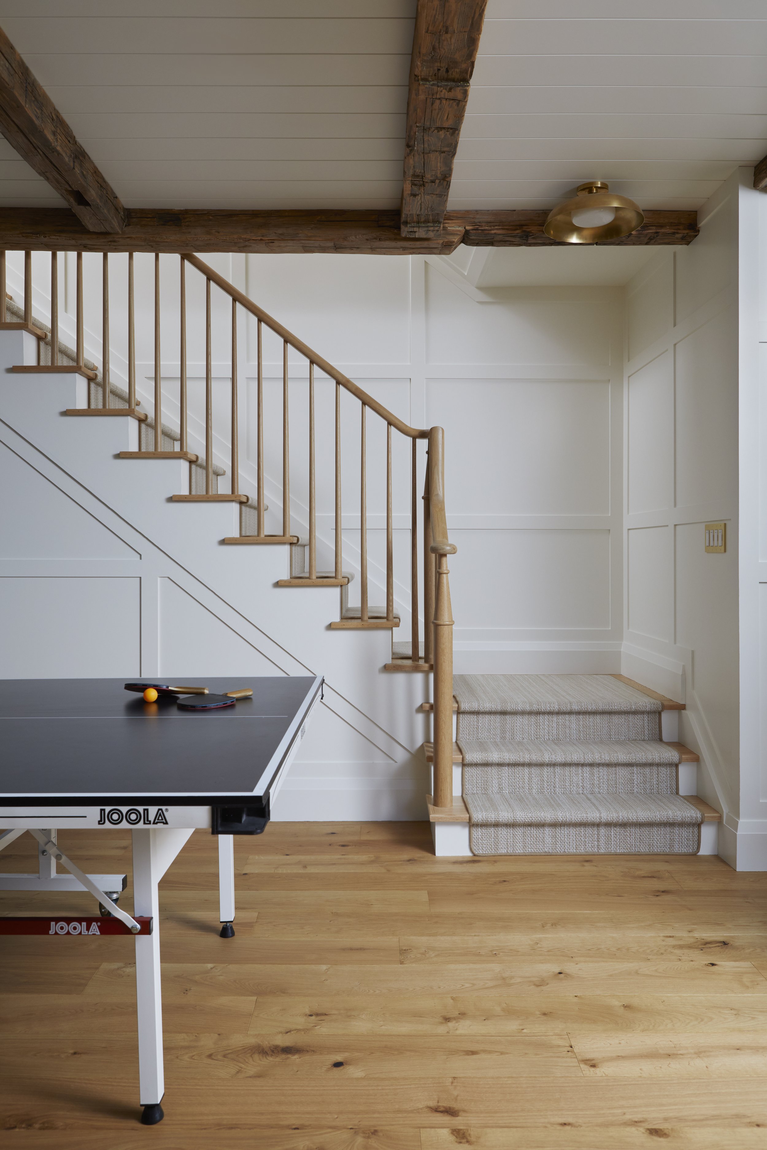Room with a ping-pong table and a staircase with wooden railing leading upstairs, wood floors, and white paneled walls.
