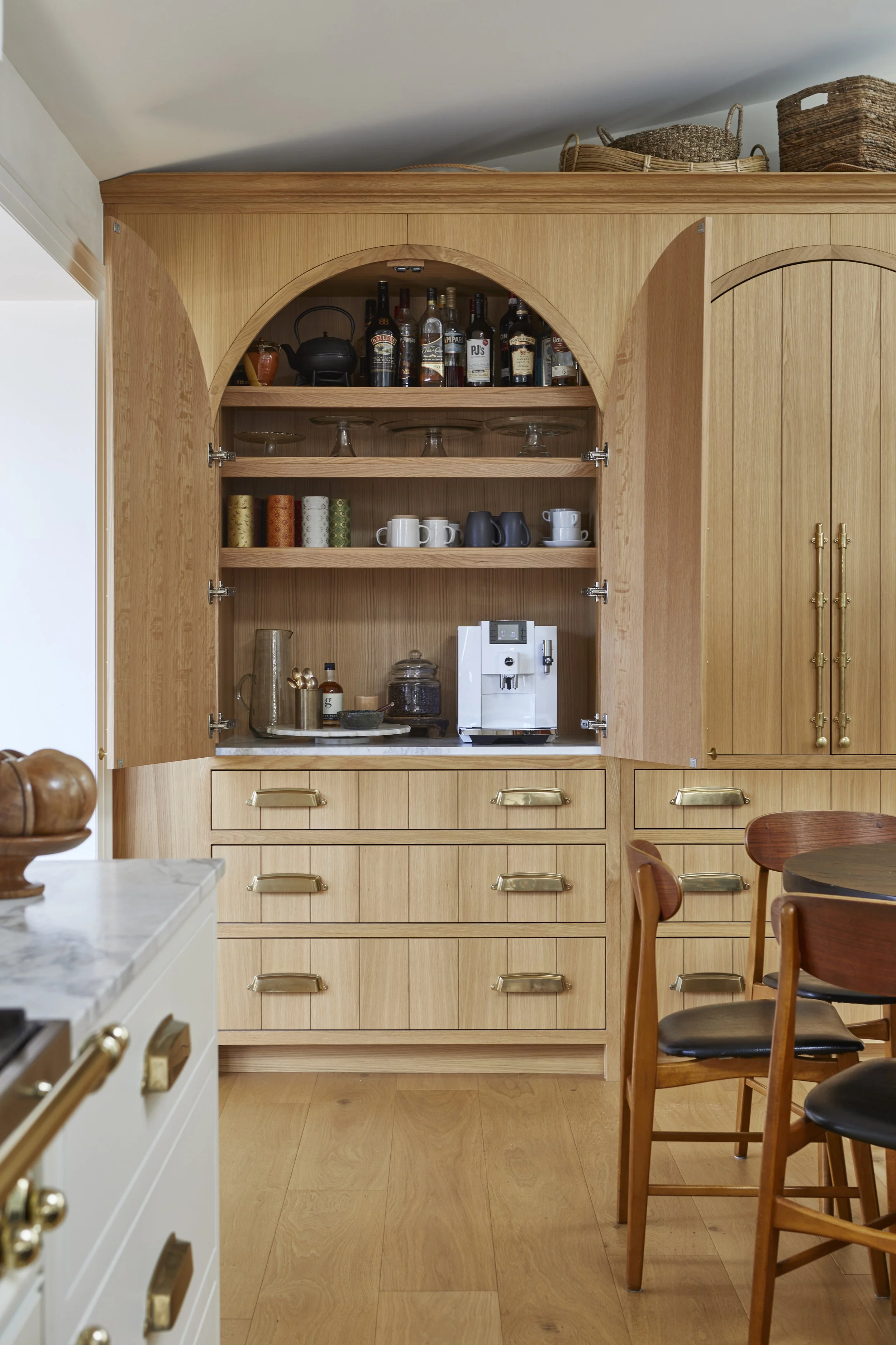 A wooden kitchen cabinet with open shelves containing alcohol bottles, coffee cups, glasses, and kitchen appliances.