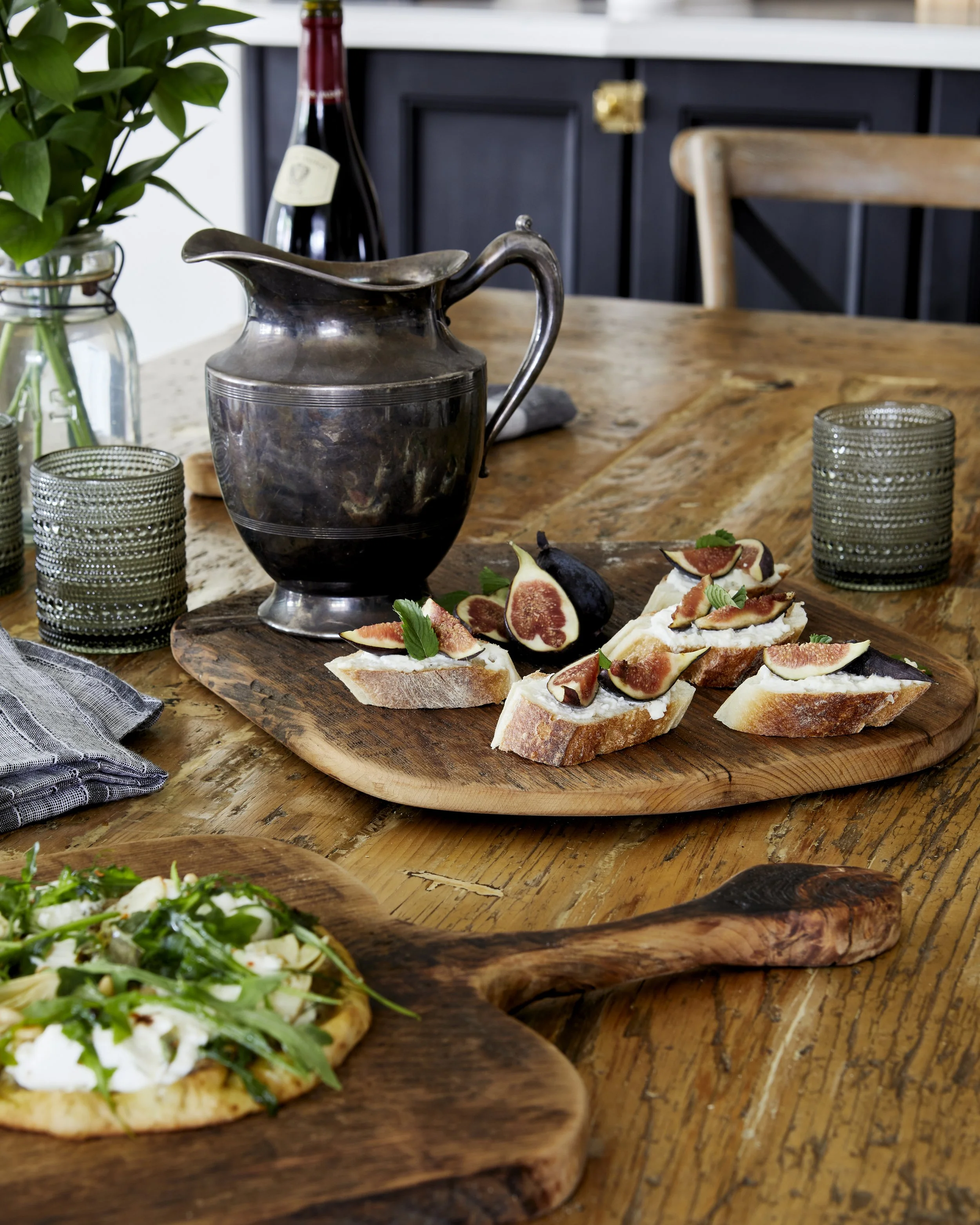 A rustic wooden dining table set with a bottle of red wine, a vintage metal pitcher, two textured green glasses, and a wooden board with fig-topped toast slices garnished with herbs. In the foreground, there's a small pizza with greens on a round woo