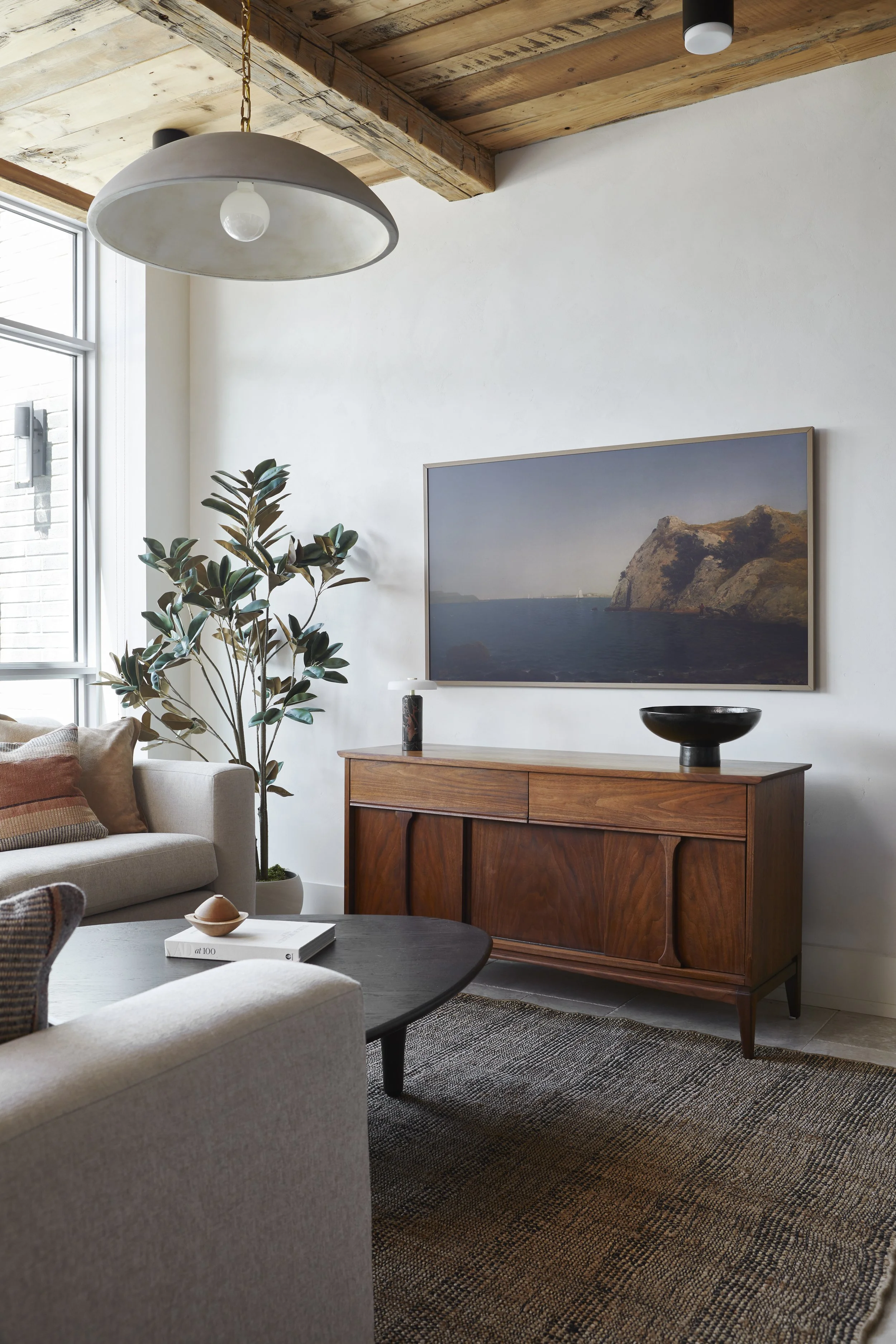 Modern living room with a beige sofa, a wooden coffee table, a tall potted plant, a white wall-mounted TV showing a landscape scene, a wooden sideboard, a black decorative bowl, and a round pendant light hanging from a wooden ceiling.
