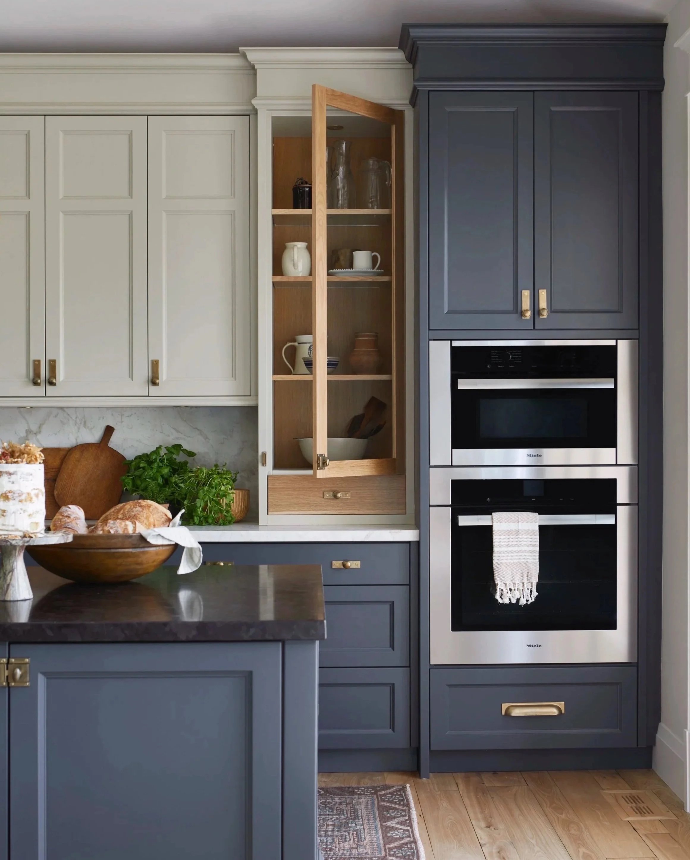 Kitchen with blue cabinetry, built-in oven, open glass cabinet, cauliflower, herbs, bread, and a black countertop.