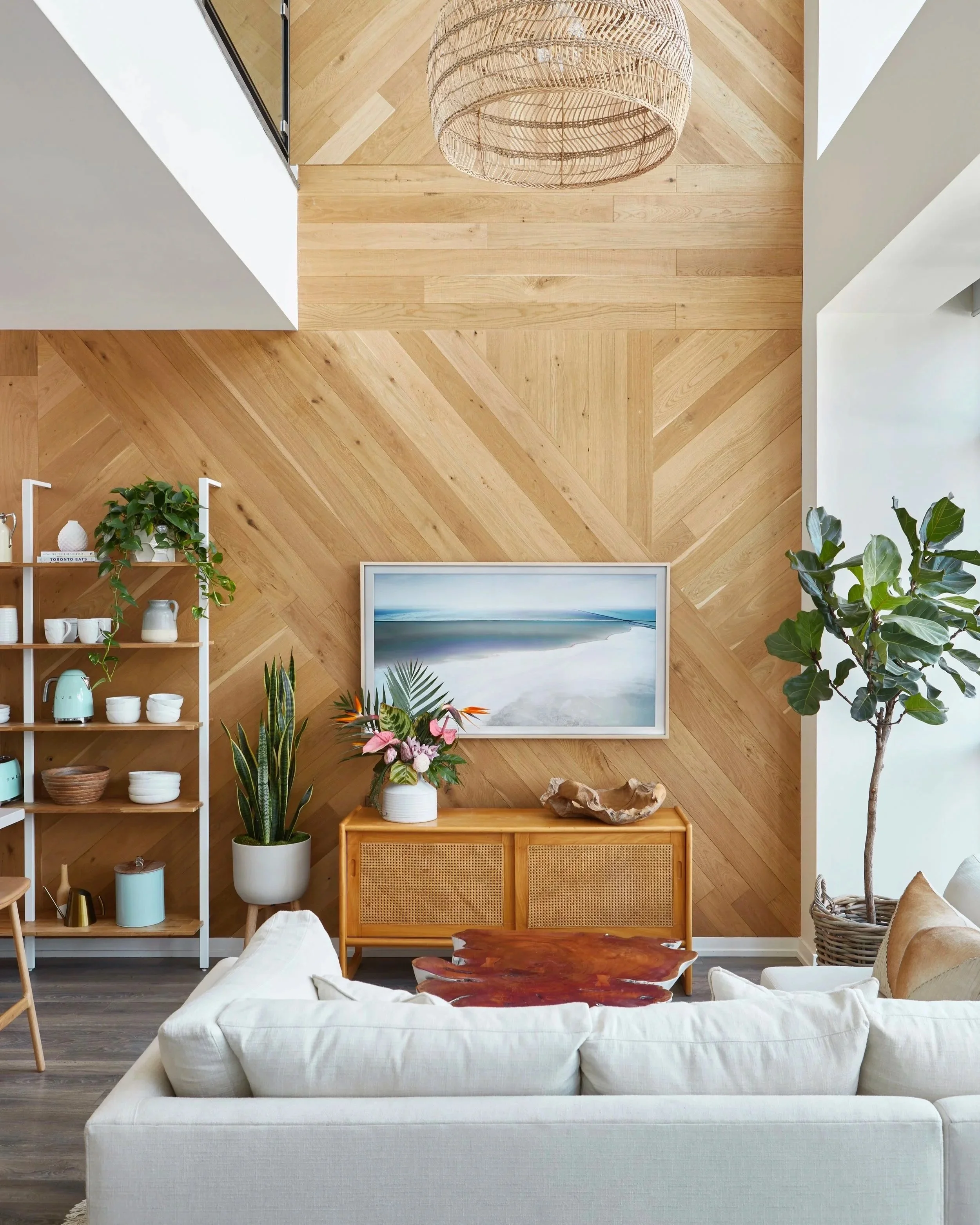 Living room with a wooden accent wall, a white sofa, and various potted plants. A framed artwork depicting a coastal scene hangs on the wall above a wooden console table.