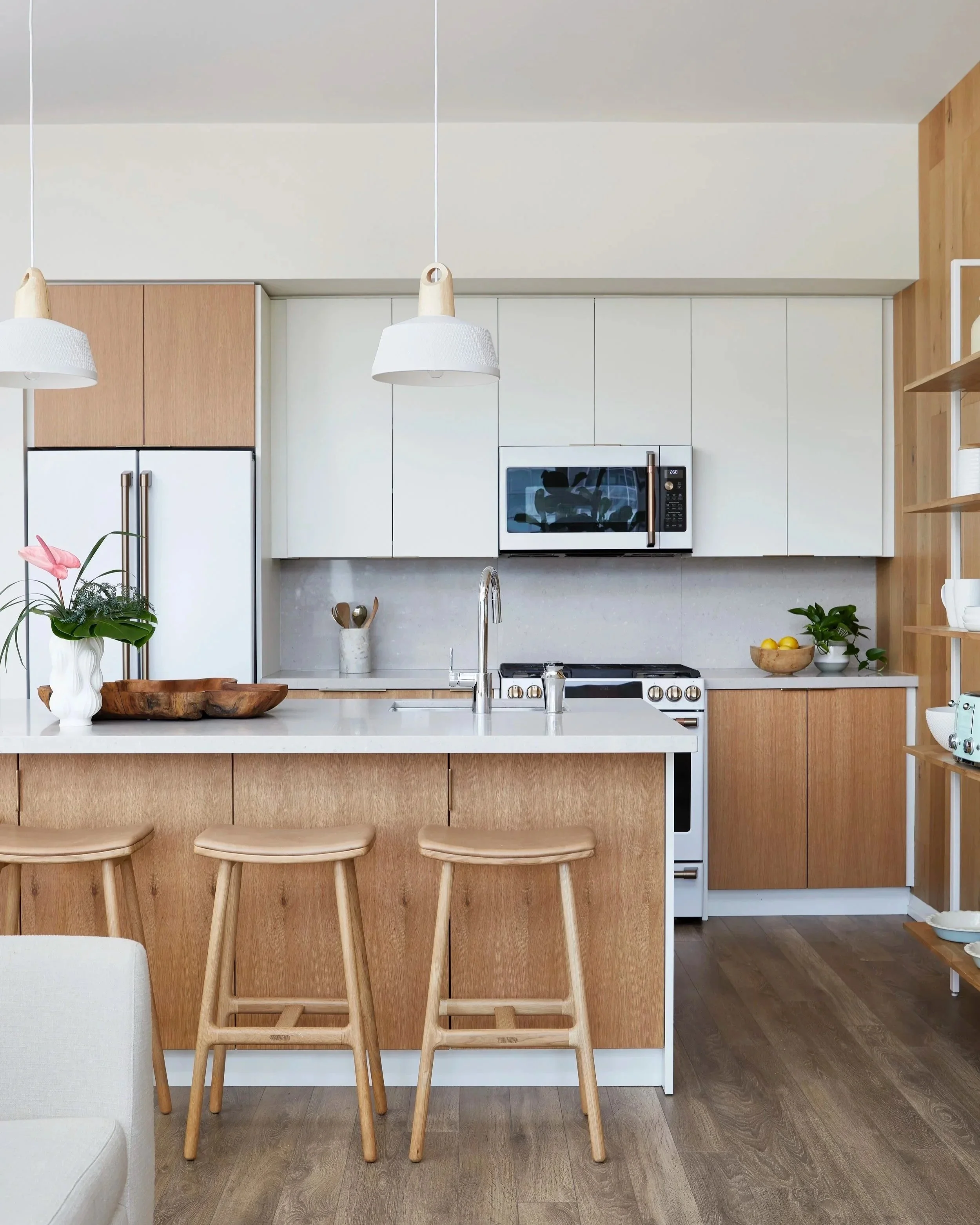 Modern kitchen with wooden cabinets, white countertops, and a kitchen island with three wooden stools. Contains a microwave, refrigerator, and various potted plants and decor.